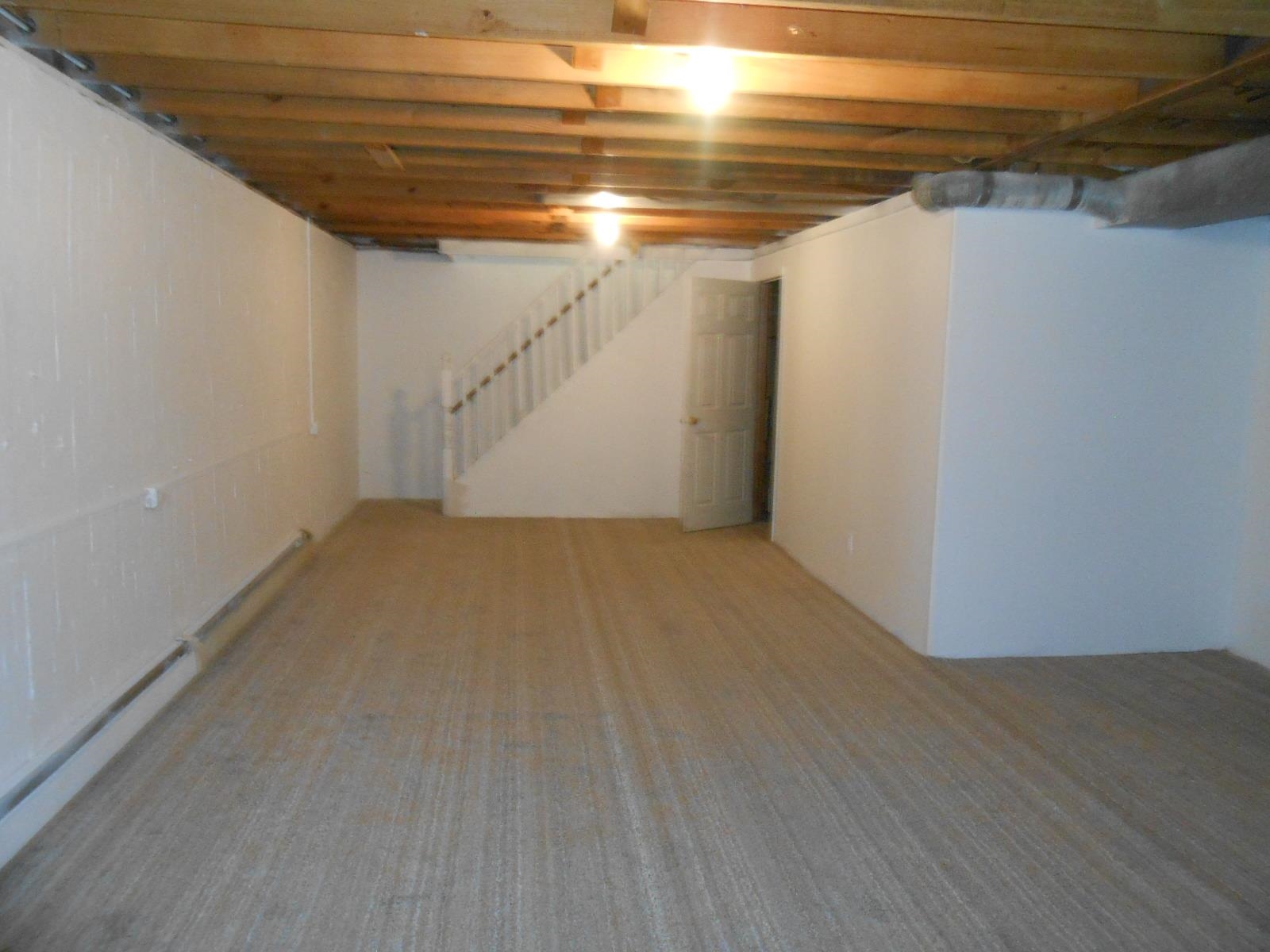 Spacious room featuring light-colored carpeting, white painted walls, exposed ceiling joists, and a partial staircase with white railing