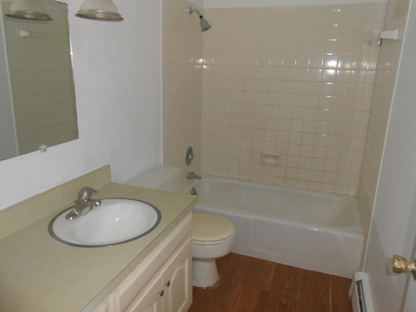 Bathroom featuring a single vanity with an integrated sink, a built-in mirror, a full bathtub with a shower, ceramic wall tiling, and wood-finish flooring