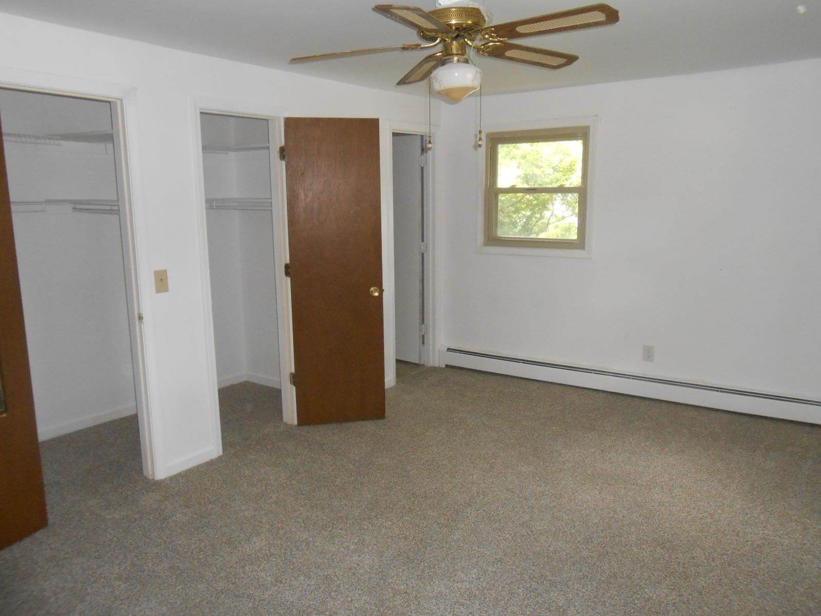 Spacious room with neutral carpeting, white walls, a ceiling fan with integrated lighting, and multiple closet storage options with wire shelving