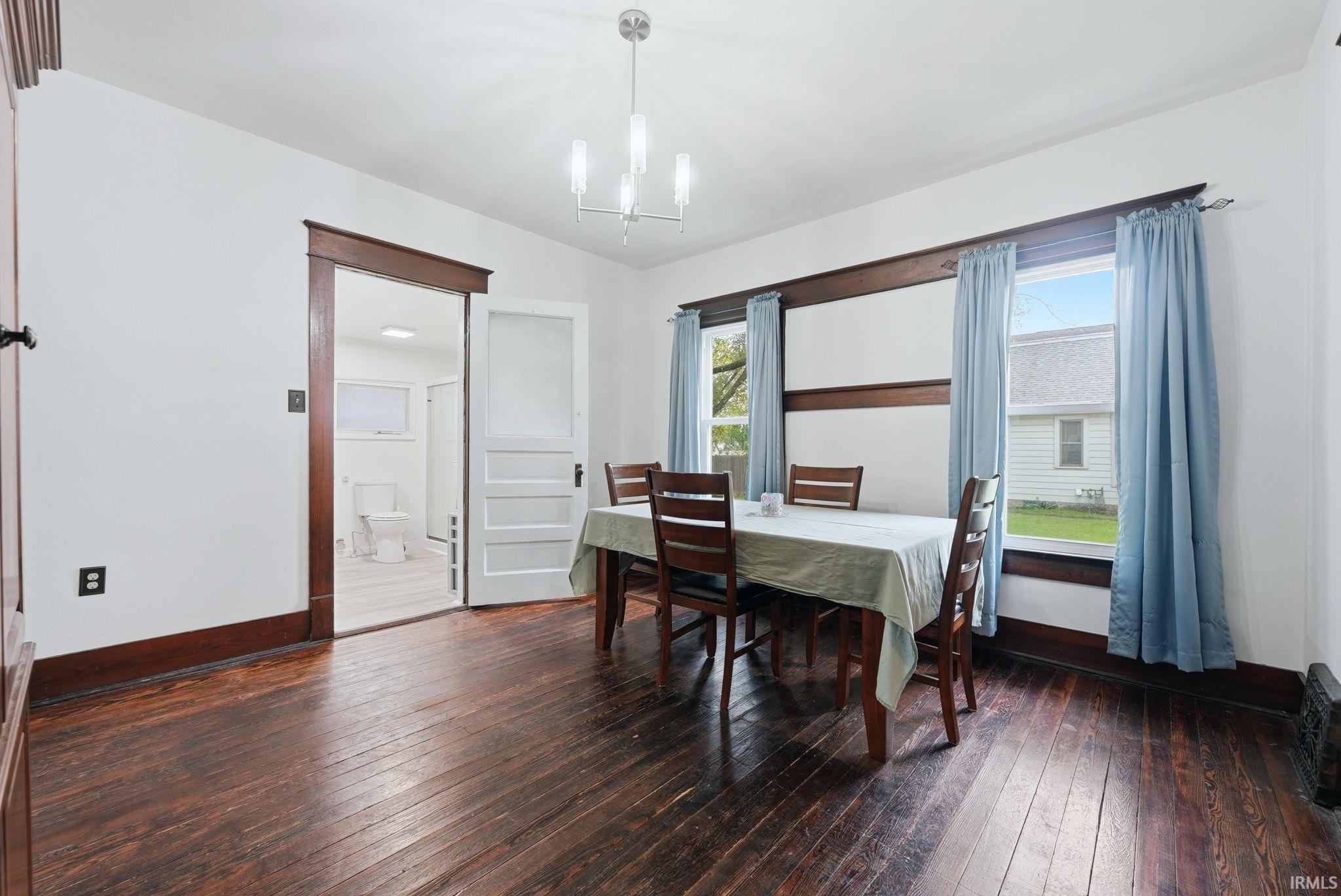 Dining area featuring dark wood-finish flooring, a contemporary chandelier, two windows with dark wood trim, and a doorway to an adjoining room
