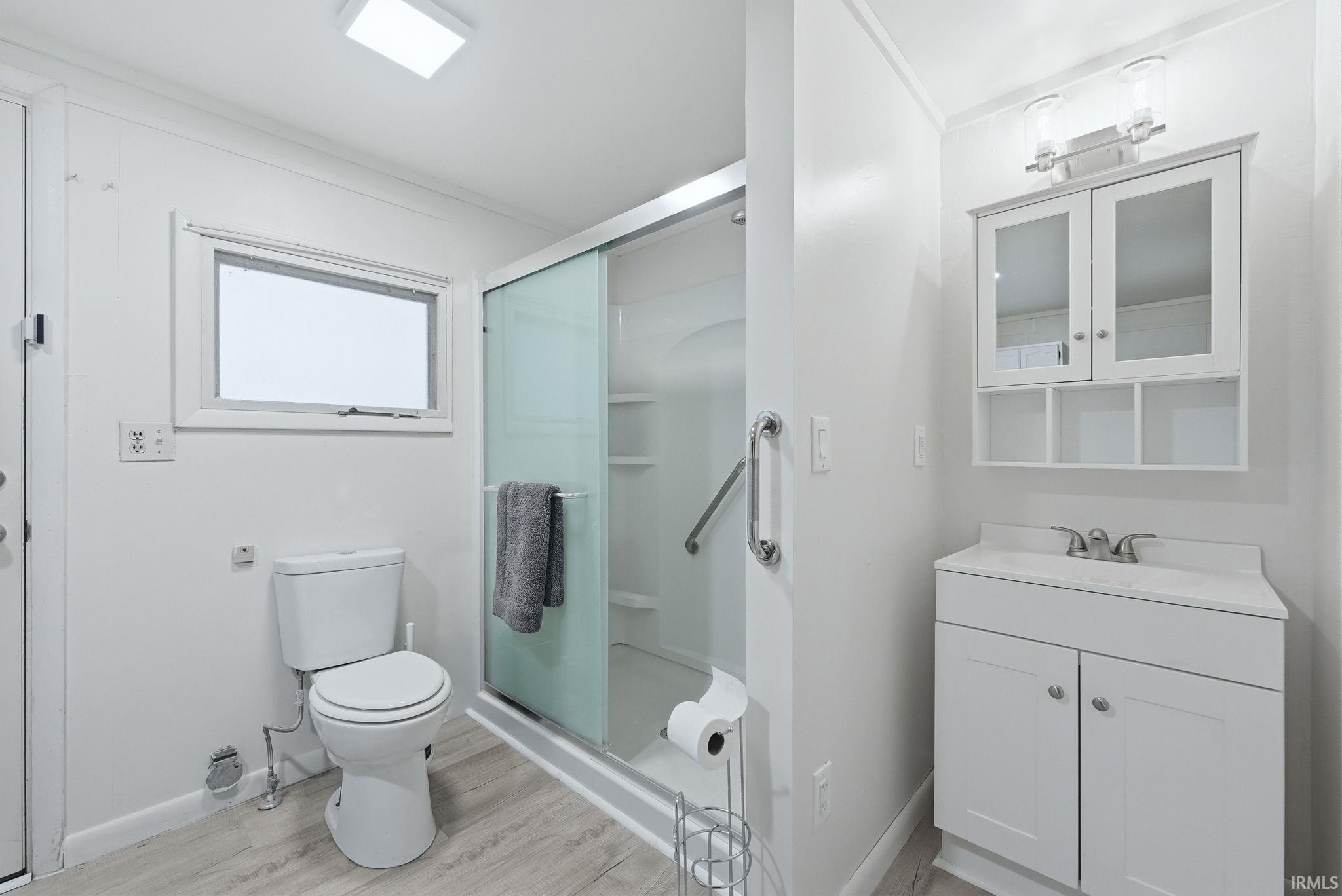 Modern bathroom featuring a sliding glass shower enclosure with grab bar, a white vanity with an integrated sink, a wall-mounted medicine cabinet with open shelving, and light wood-finish flooring