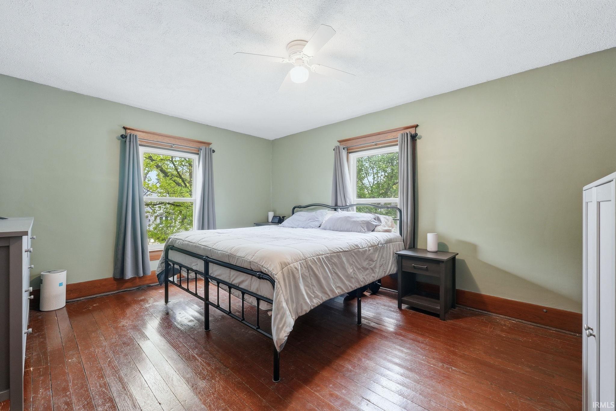 Bedroom featuring wood-finish flooring, two window openings with natural wood trim, and a ceiling fan with integrated lighting