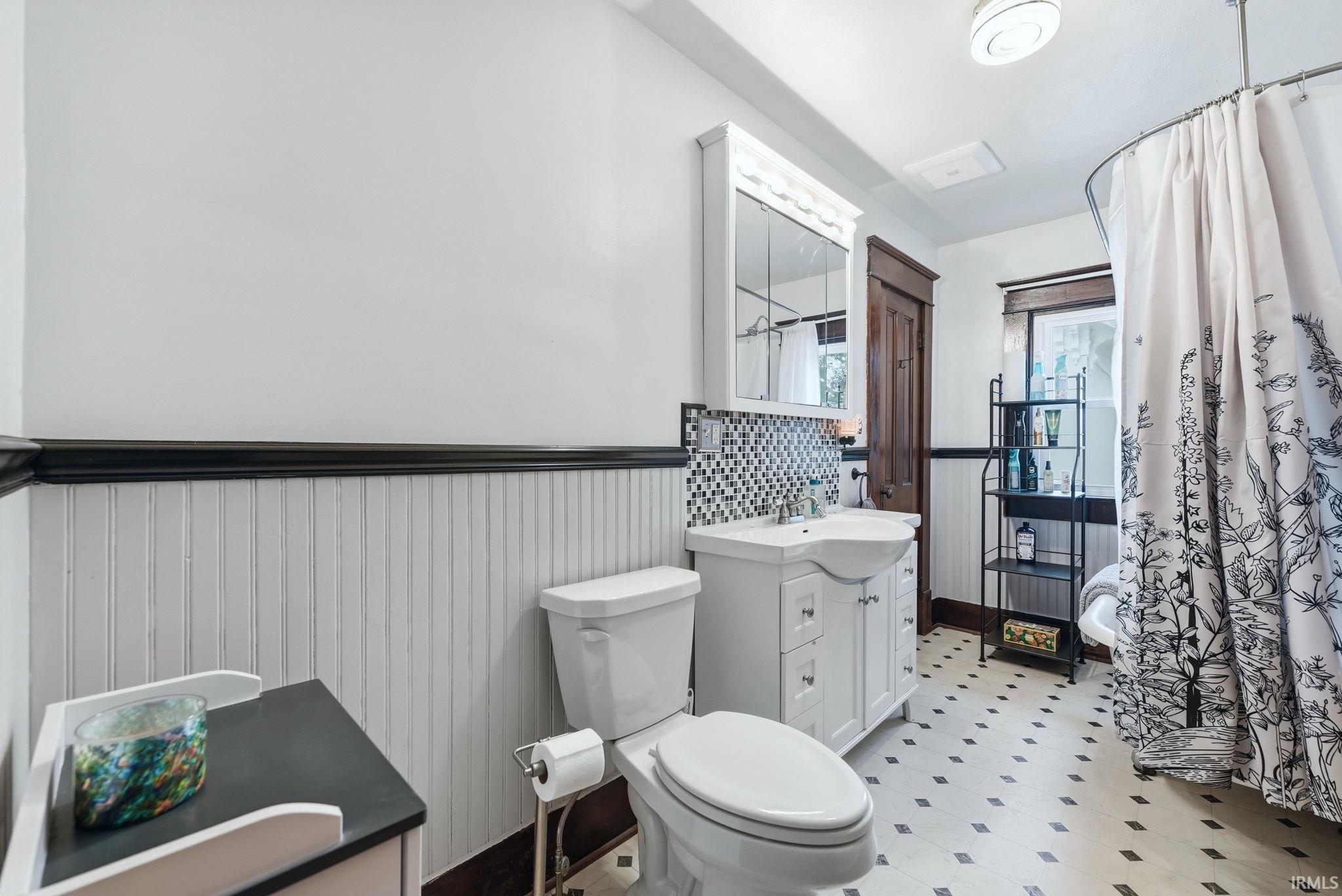 Bathroom featuring beadboard wainscoting, a white vanity with an integrated sink, a mosaic tile backsplash, a toilet, and patterned floor tiles