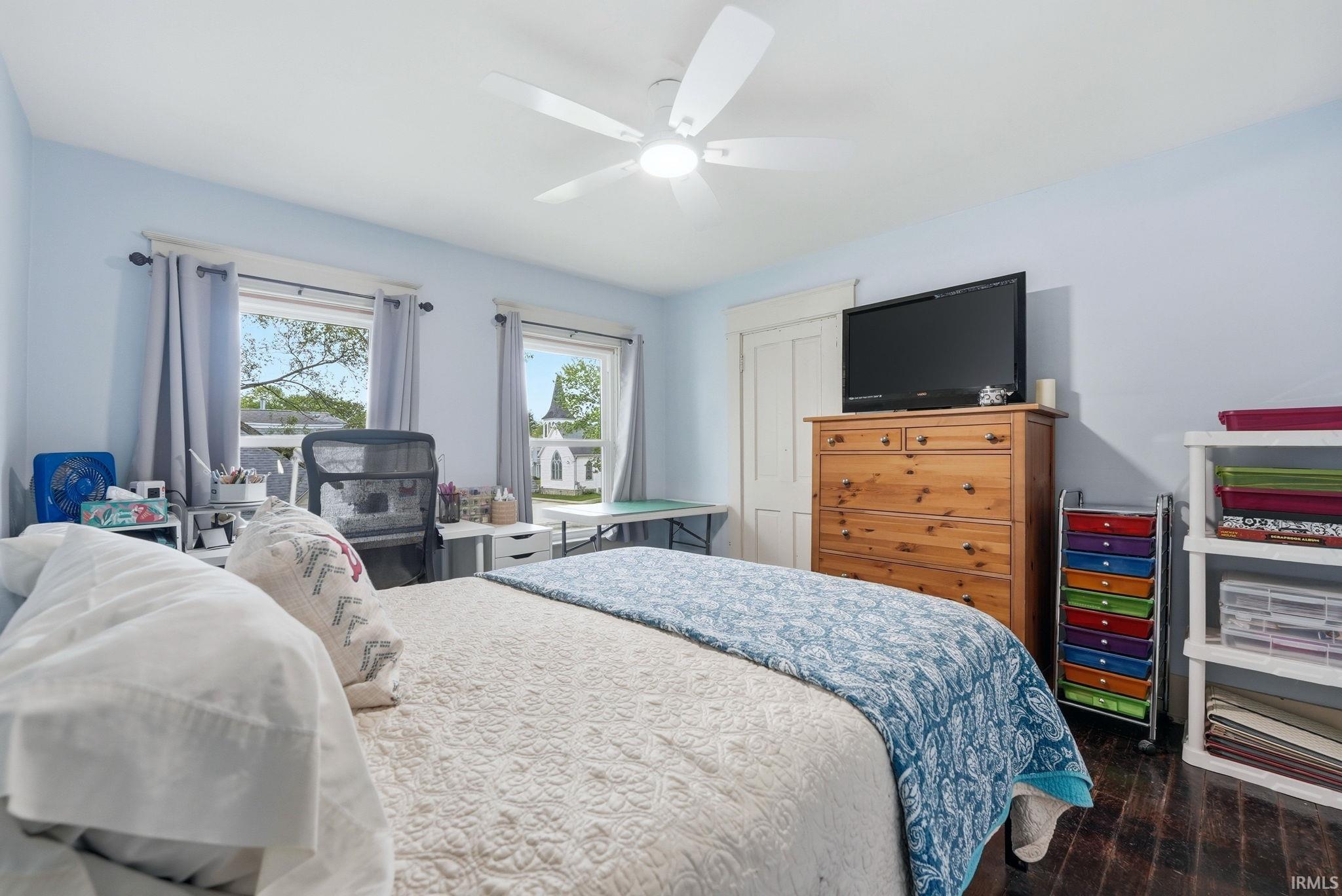 Bedroom featuring two windows, a ceiling fan, light blue walls, dark wood-finish flooring, and a white paneled door
