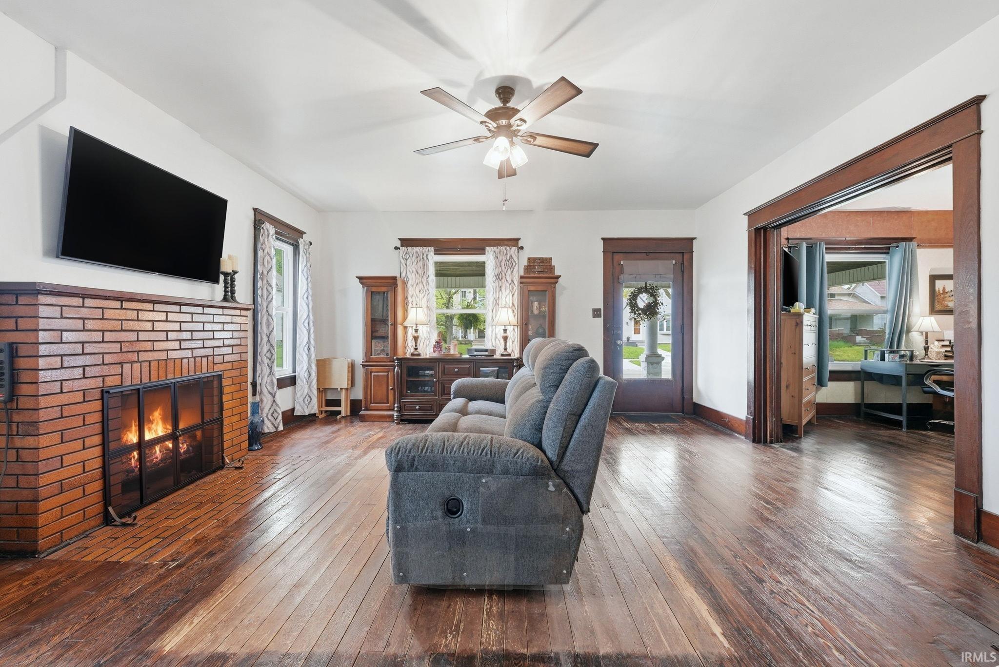 Living area featuring a brick fireplace, wood-finish flooring, a ceiling fan, and painted white walls