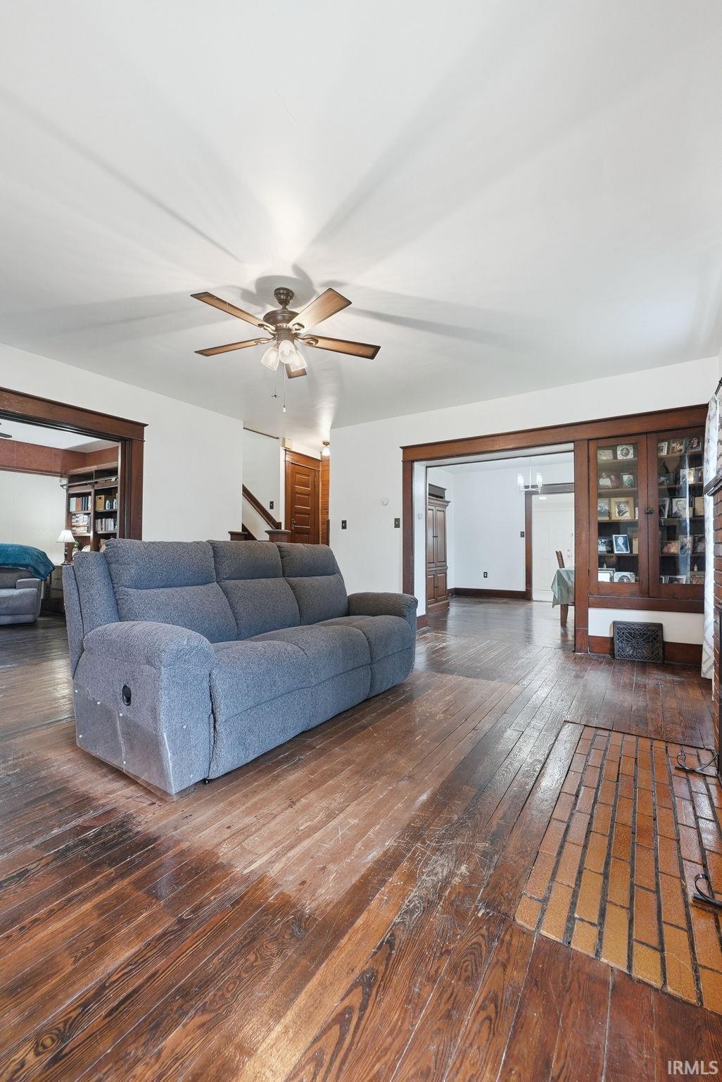 Spacious living area featuring wood-finish flooring, a ceiling fan with integrated lighting, and a brick-tiled hearth