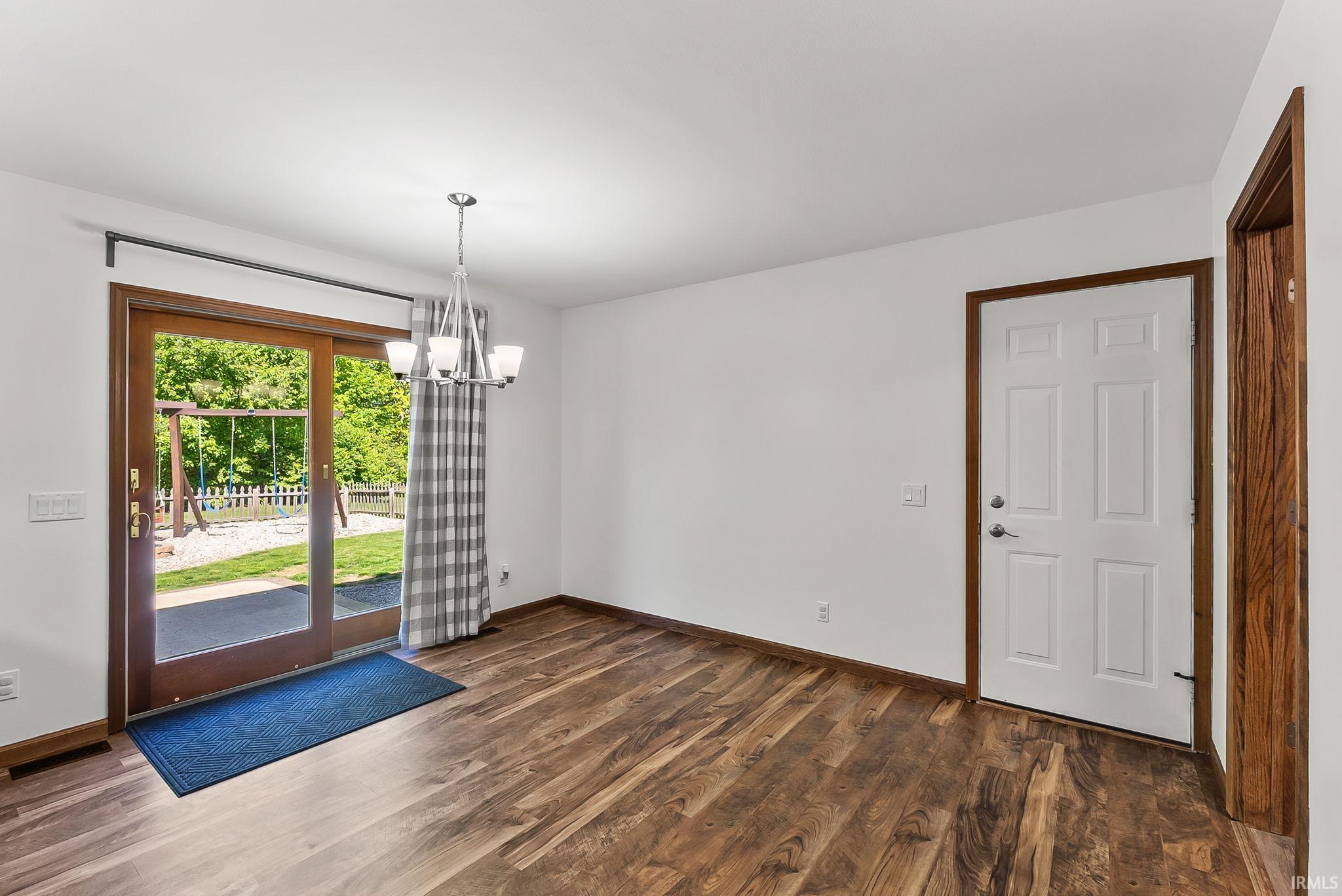 Spacious room featuring wood-finish flooring, a contemporary chandelier, and a white six-panel entry door with wood trim