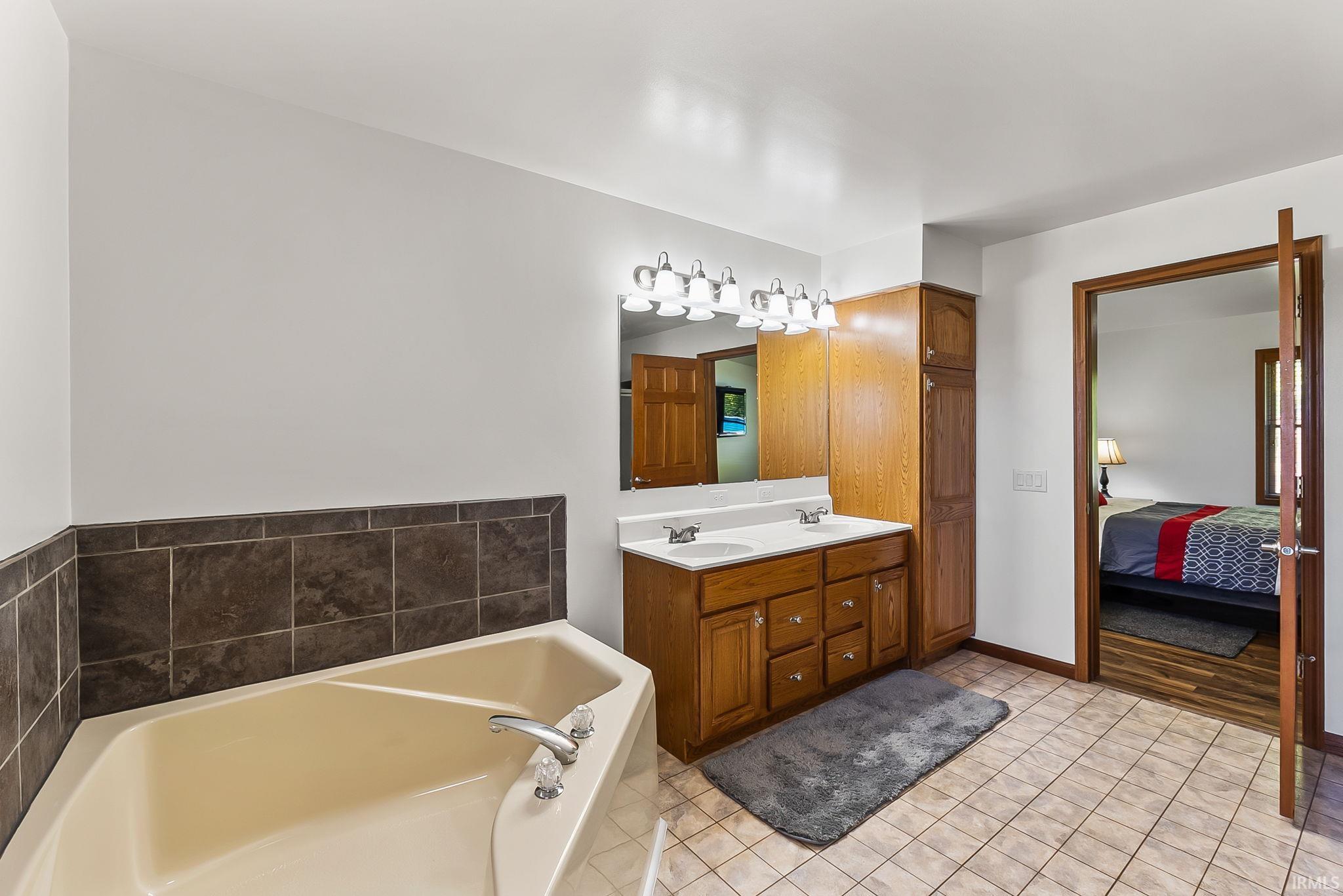 Bathroom featuring an angled soaking tub with dark tile surround, a dual vanity with wood-finish cabinetry and integrated sinks, and tiled flooring
