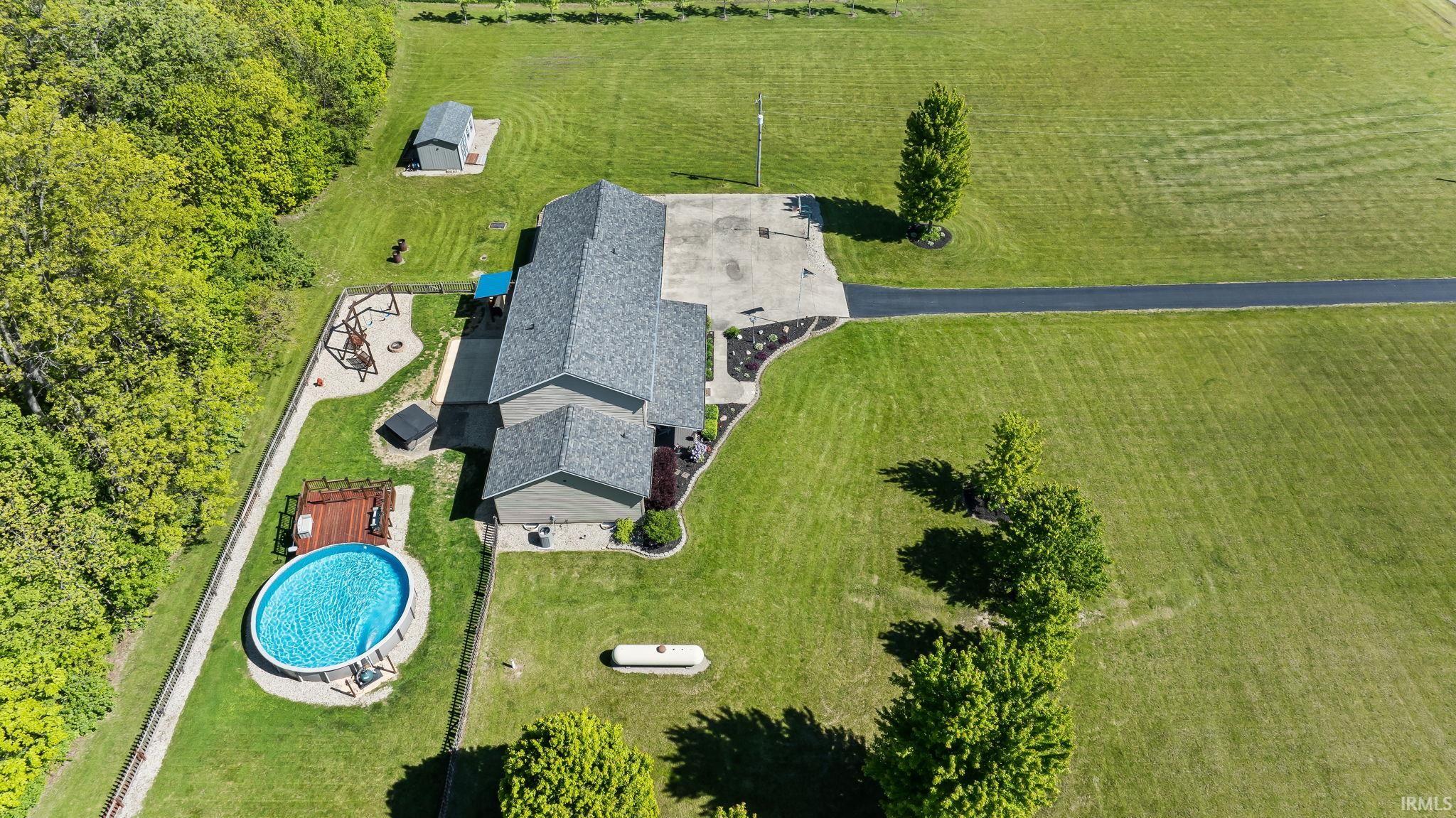Expansive property featuring a grey-shingled residence with a large concrete patio, a dedicated above-ground pool area with decking, a separate storage shed, and a paved driveway surrounded by lush green lawns