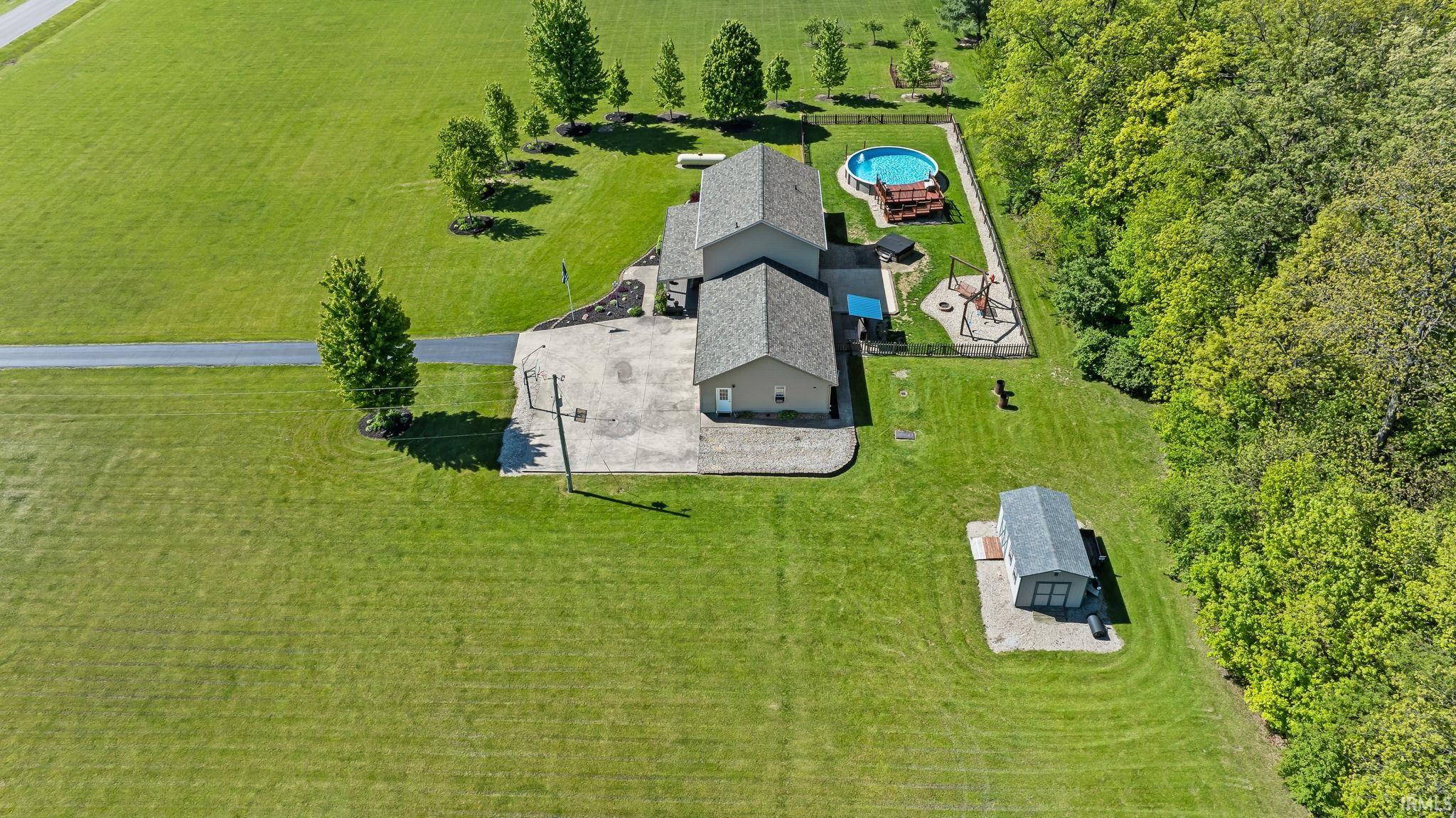 Expansive property featuring a residence with a light gray roof, a detached shed, a paved driveway, a circular above-ground pool, and a wooded border