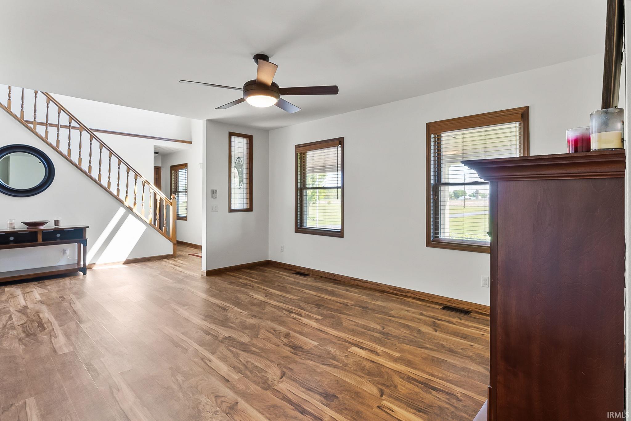 Open living area featuring wood-finish flooring, a ceiling fan with integrated lighting, two windows with wood trim, and a white staircase with wood railing