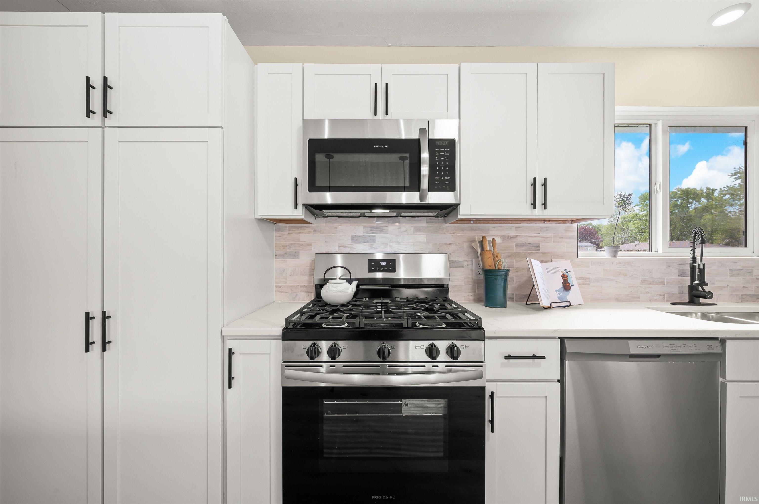 Modern kitchen featuring white shaker cabinetry, stainless steel appliances, light stone backsplash, white countertops, and a window above the sink