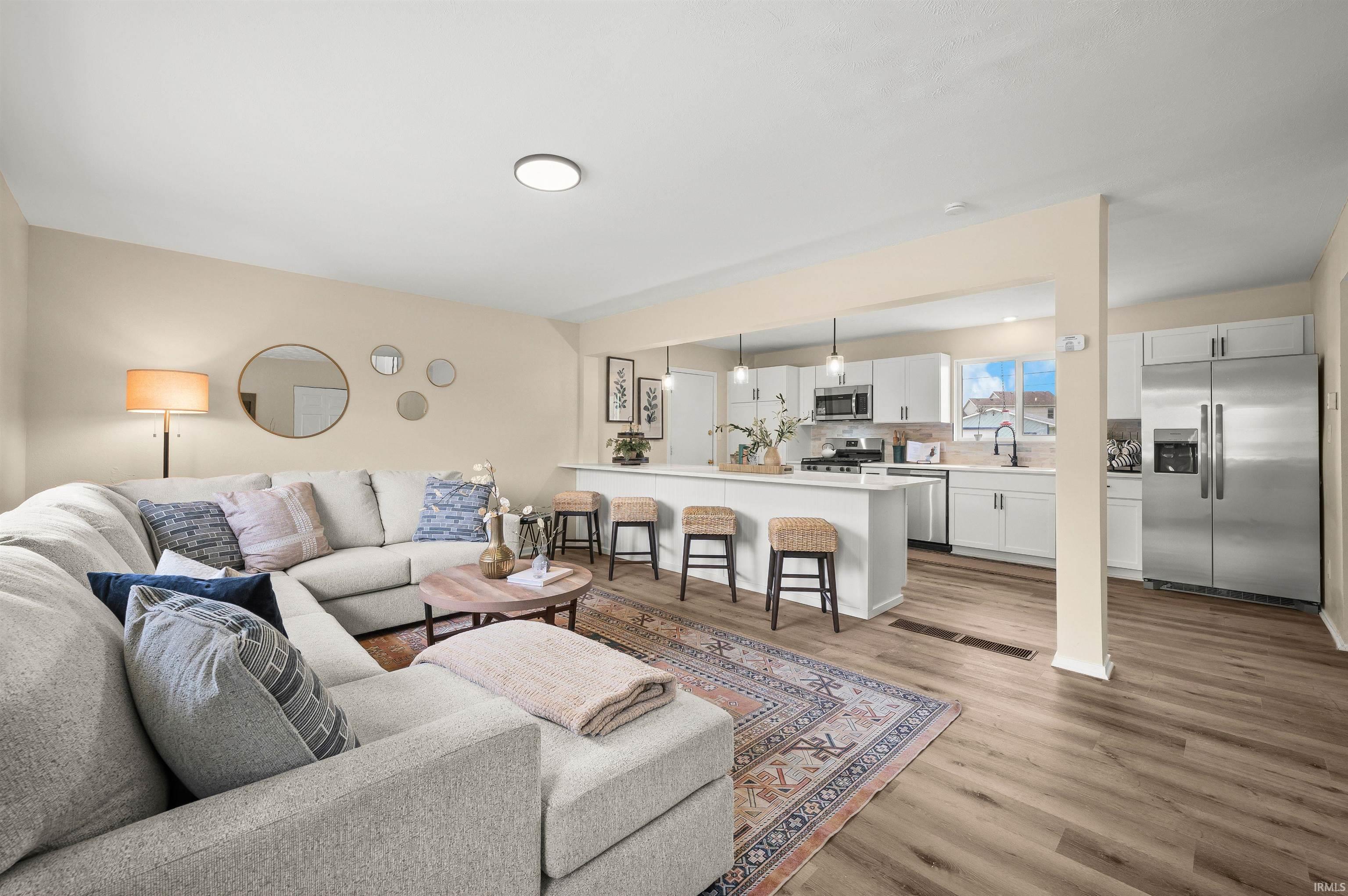 Open-concept living area featuring wood-finish flooring, a kitchen island with seating, pendant lighting, stainless steel appliances, and white cabinetry