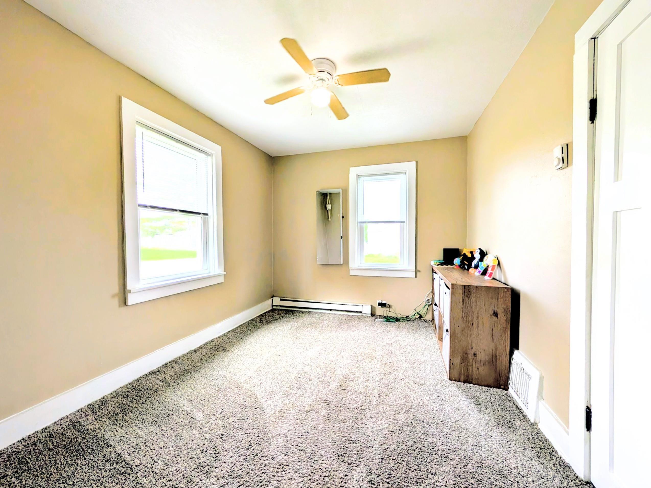 Carpeted interior space featuring two windows with white trim, a ceiling fan with light fixture, baseboard heating element, and light tan wall finishes