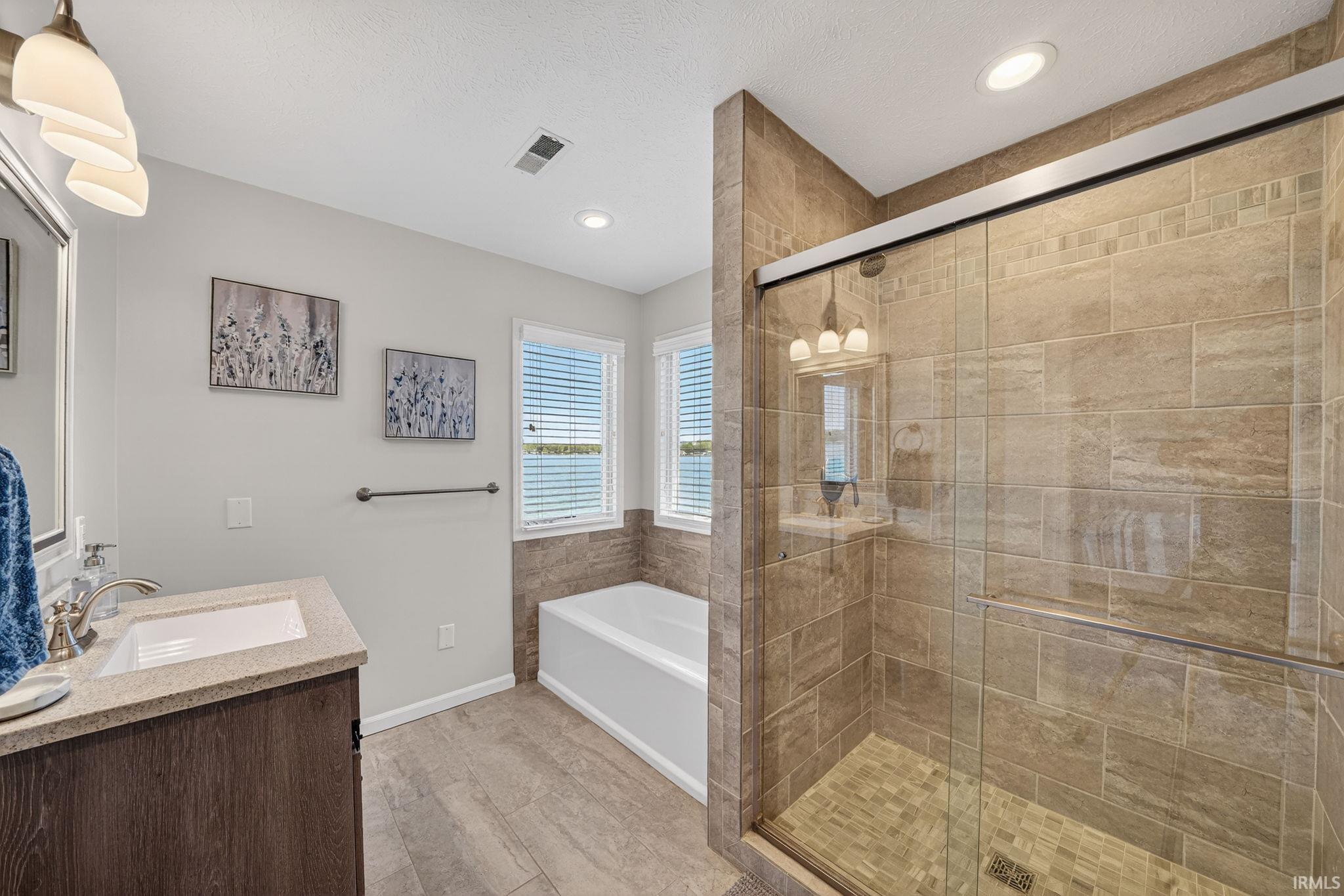 Spacious bathroom featuring a glass-enclosed shower with tile surround, a built-in soaking tub, a single vanity with stone-finish countertop, and wood-finish flooring