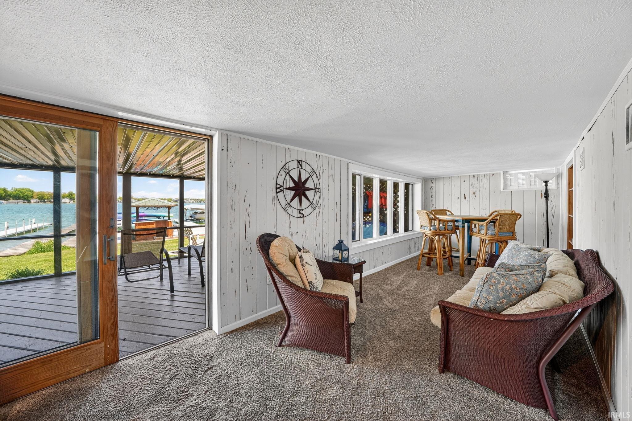 Spacious sunroom featuring sliding glass doors opening to a covered deck, durable carpet flooring, textured white wall paneling, and a horizontal window with multiple panes
