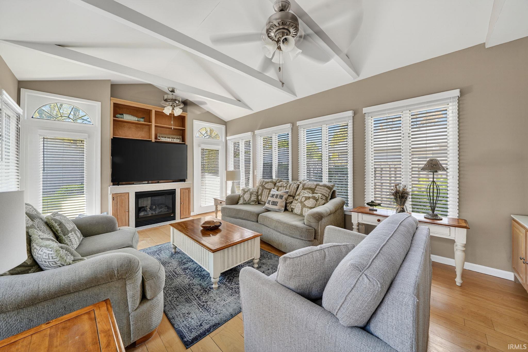 Vaulted ceiling with exposed white beams and ceiling fans, wood-finish flooring, built-in entertainment center with fireplace, multiple windows with blinds, and transom windows above French doors