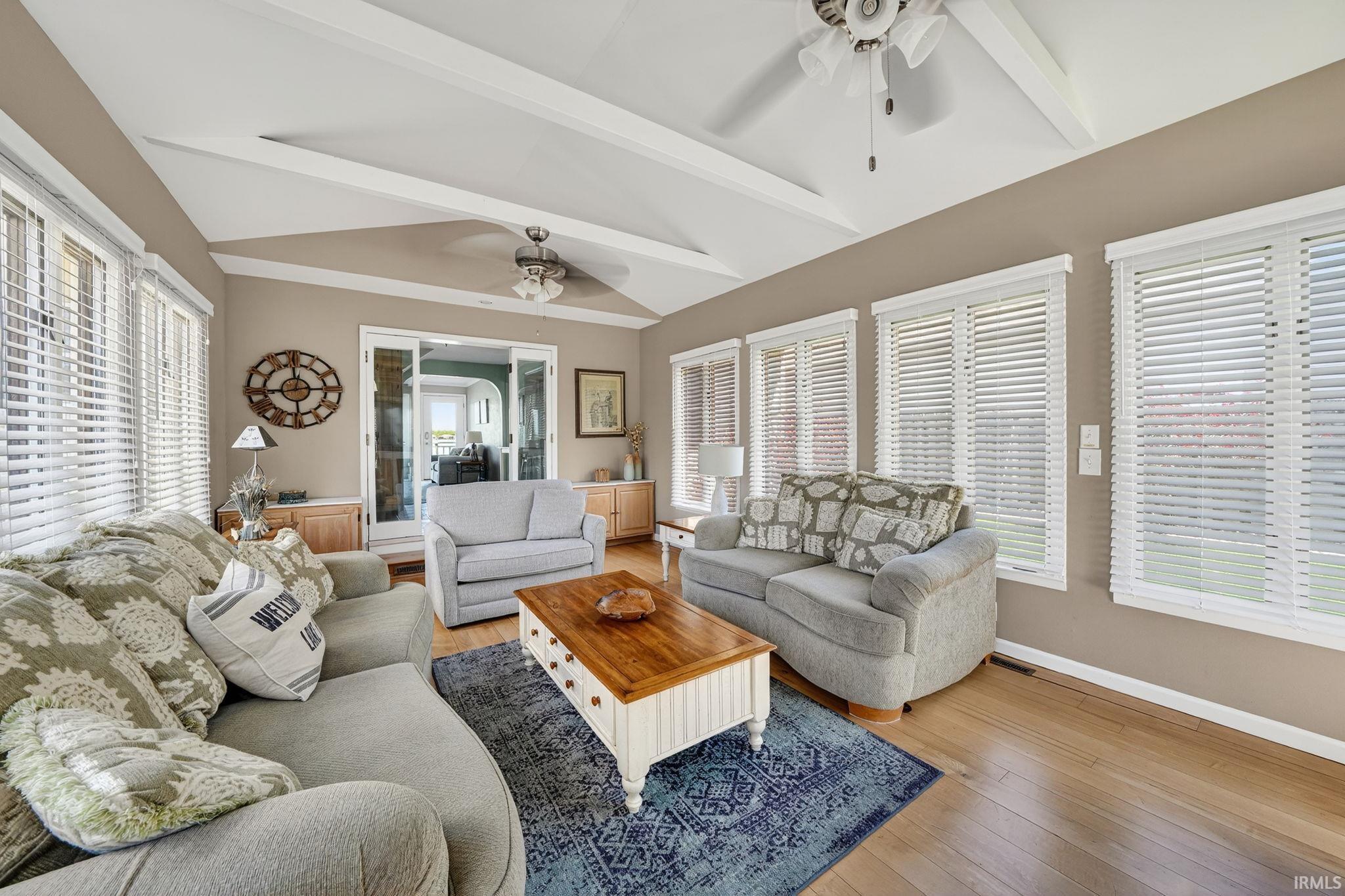Living space featuring vaulted ceilings with exposed beams, wood-finish flooring, and multiple windows with white blinds