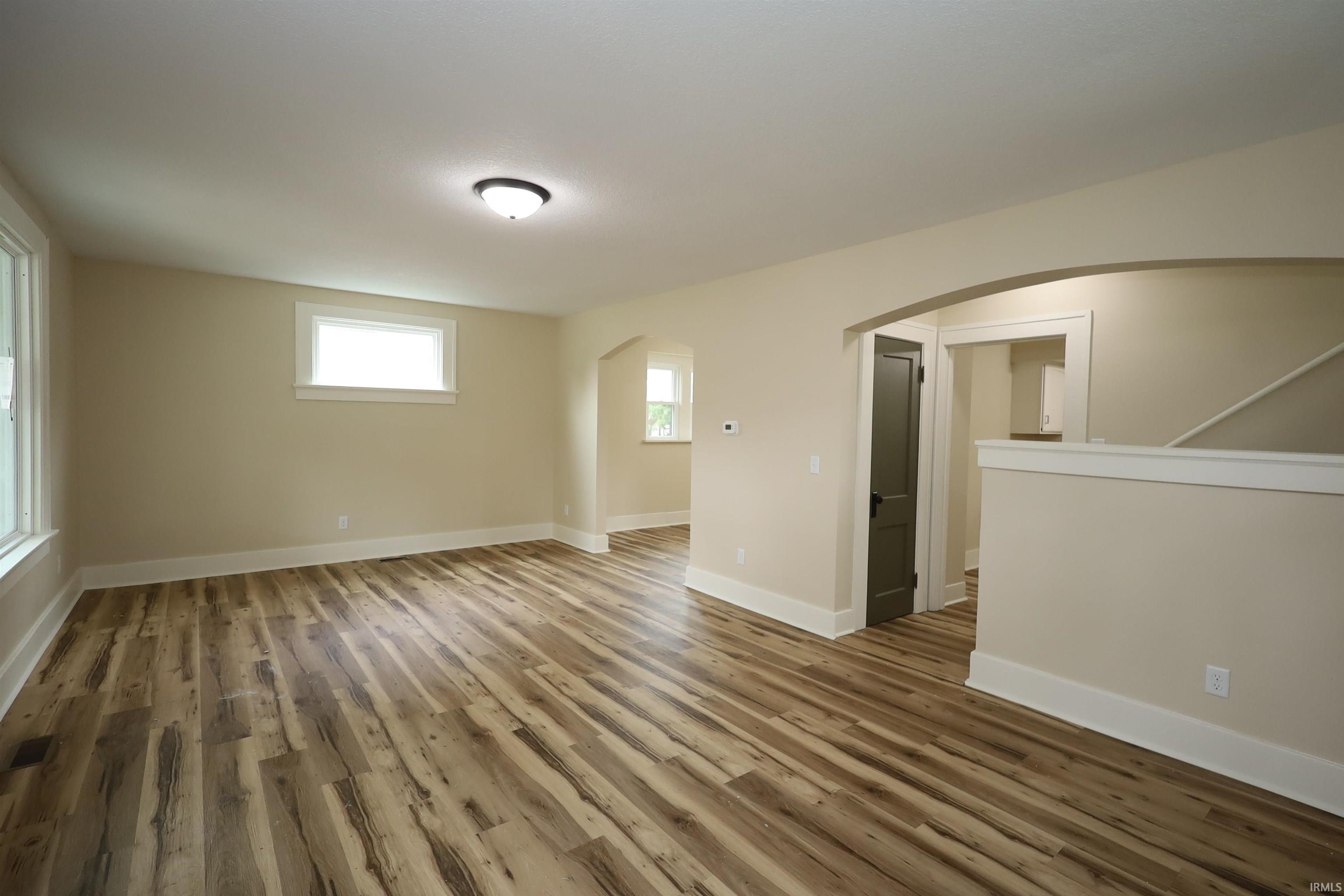 Spacious room featuring wood-finish flooring, two windows with white trim, a flush-mount ceiling light fixture, and an arched doorway