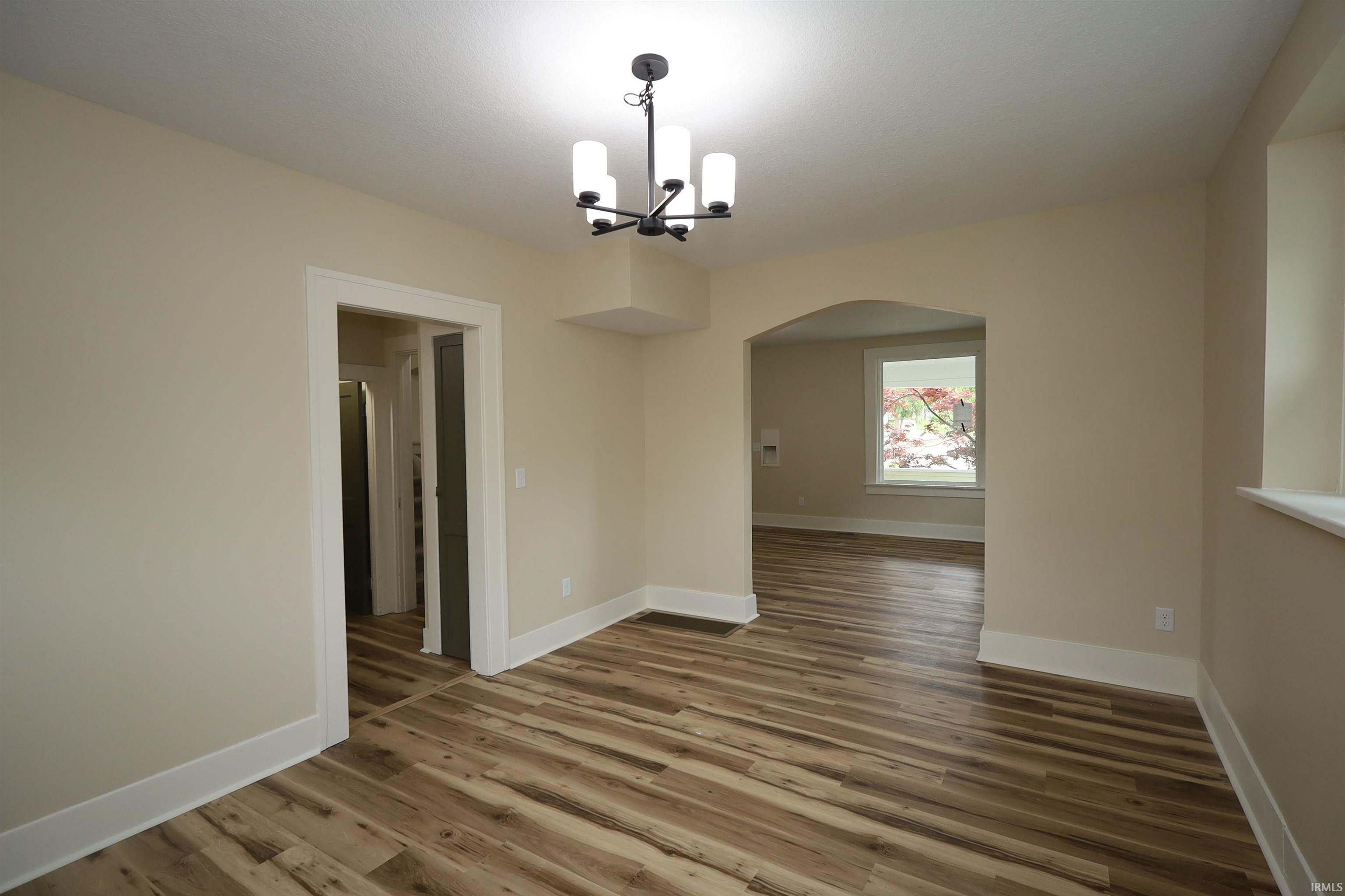 Interior space featuring wood-finish flooring, a modern black chandelier, neutral wall paint, white trim, and an arched doorway