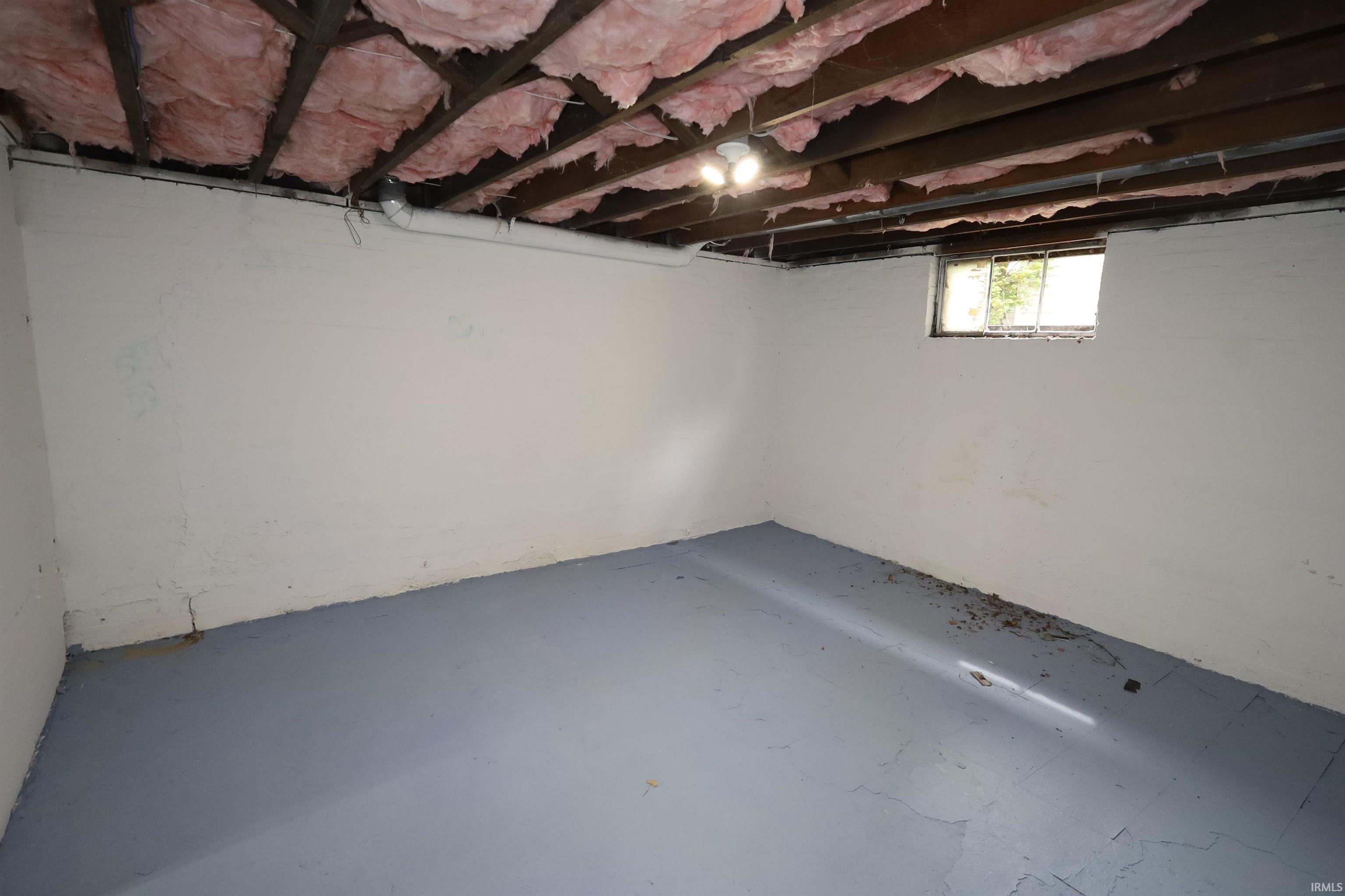 Lower-level room featuring exposed ceiling joists with insulation, a painted concrete floor, and a single window offering exterior light