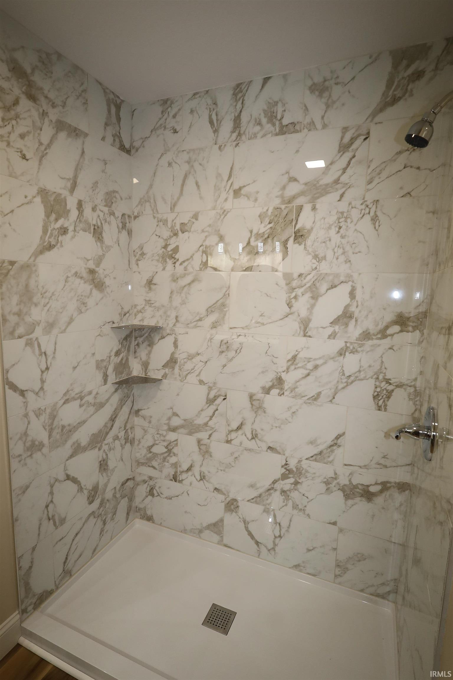 Shower enclosure featuring white and grey marbled wall tiling, a built-in corner shelf, a white shower pan with a square drain, and a chrome showerhead and control handle