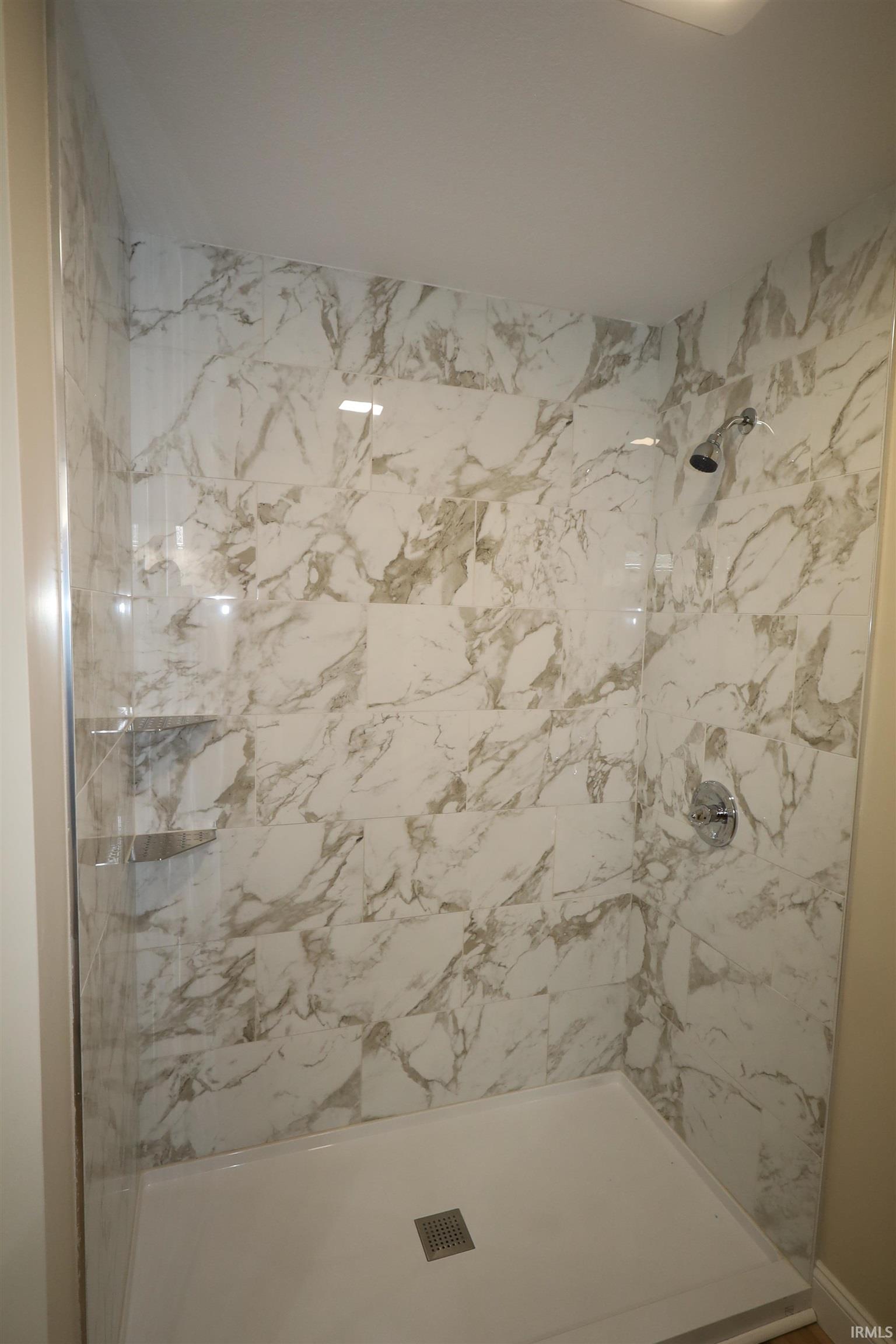 Shower enclosure featuring white marble-look tile, a white shower pan, and a chrome shower head and controls
