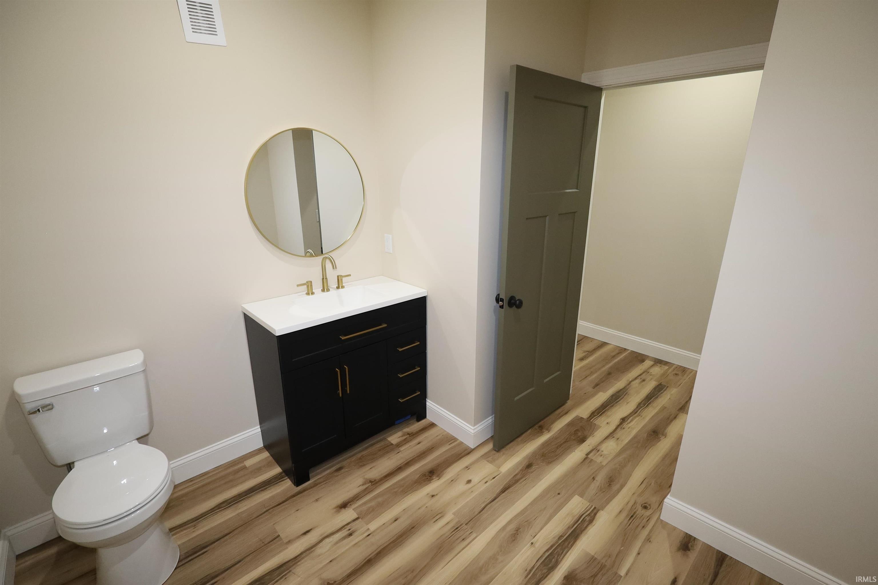 Contemporary bathroom featuring a black vanity with a white countertop, gold-finish hardware, a round gold-frame mirror, and wood-finish flooring