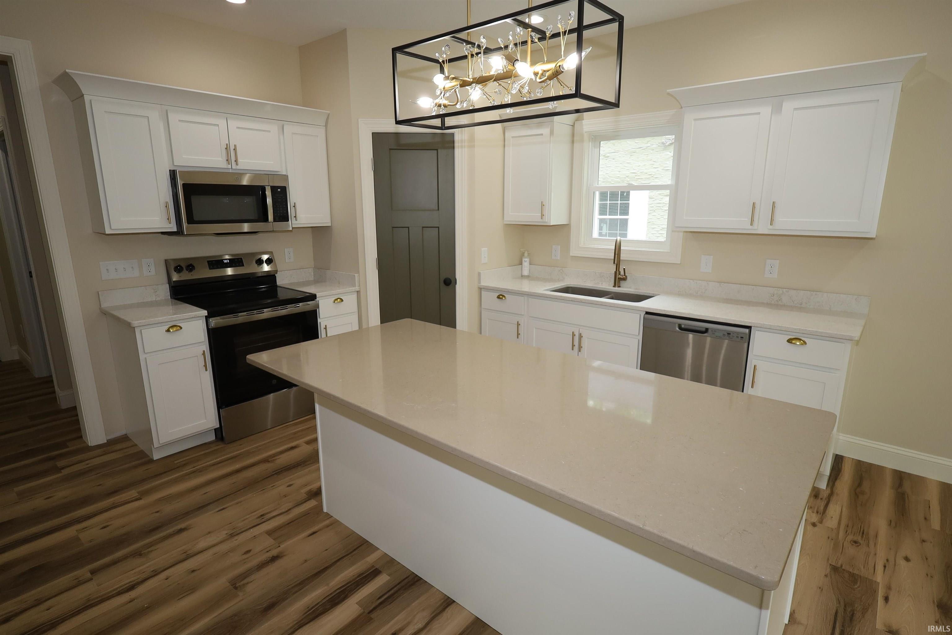 Contemporary kitchen featuring a central island with a solid surface countertop, white shaker-style cabinetry, stainless steel appliances, wood-finish flooring, and a modern rectangular chandelier