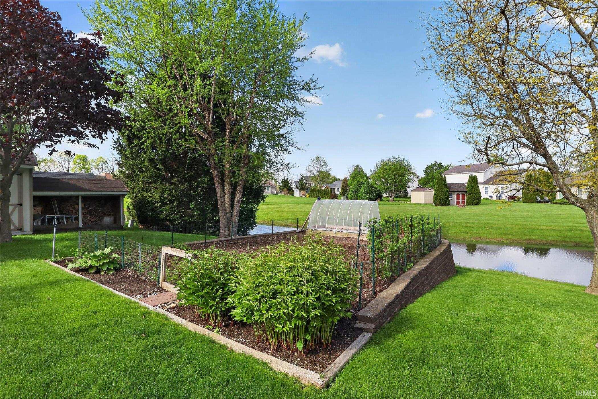 Expansive backyard featuring a manicured lawn, a large raised garden bed with a greenhouse structure, a retaining wall, and mature trees