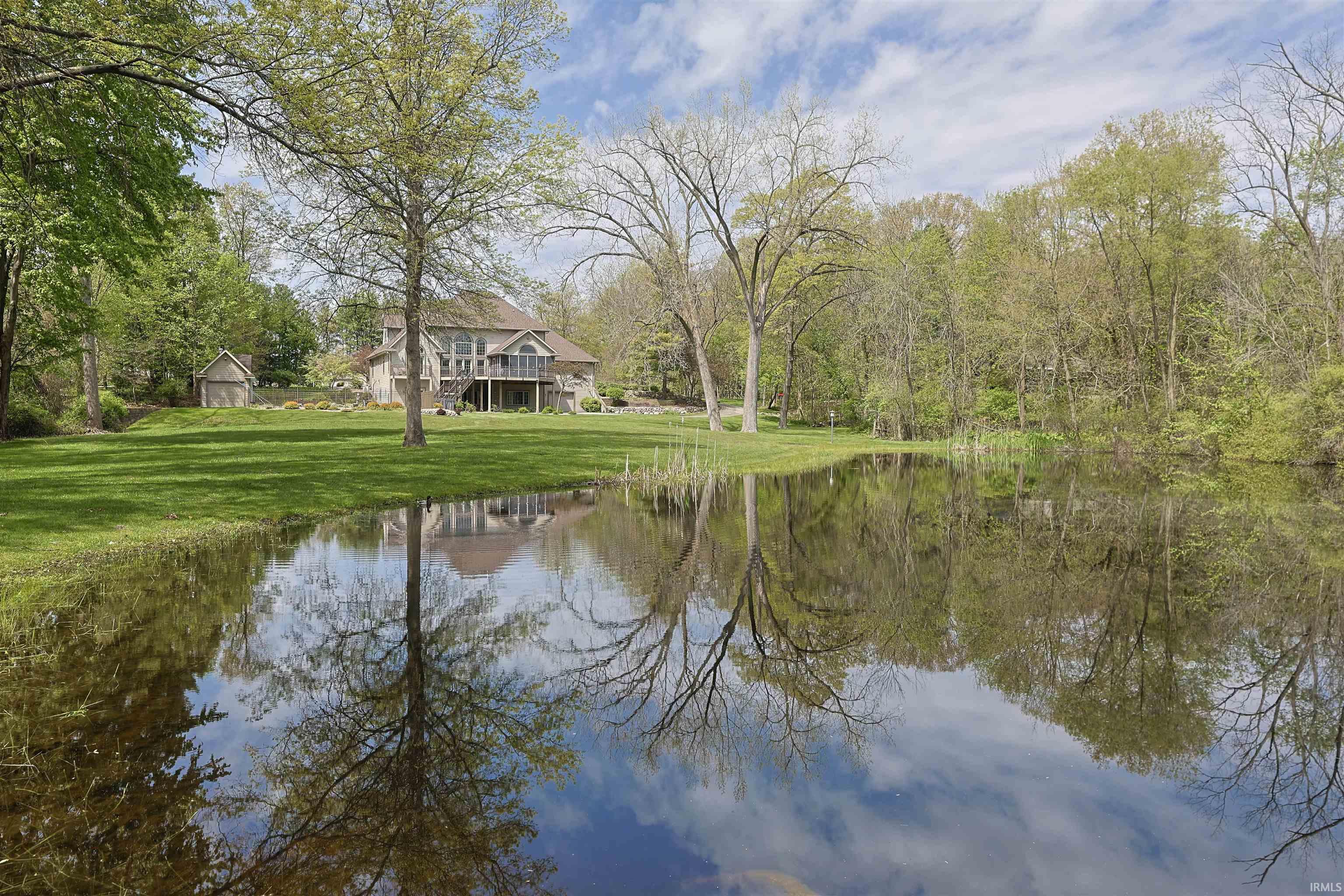 Expansive pond reflecting a treed landscape and cloudy sky, with a residential structure visible beyond a manicured lawn