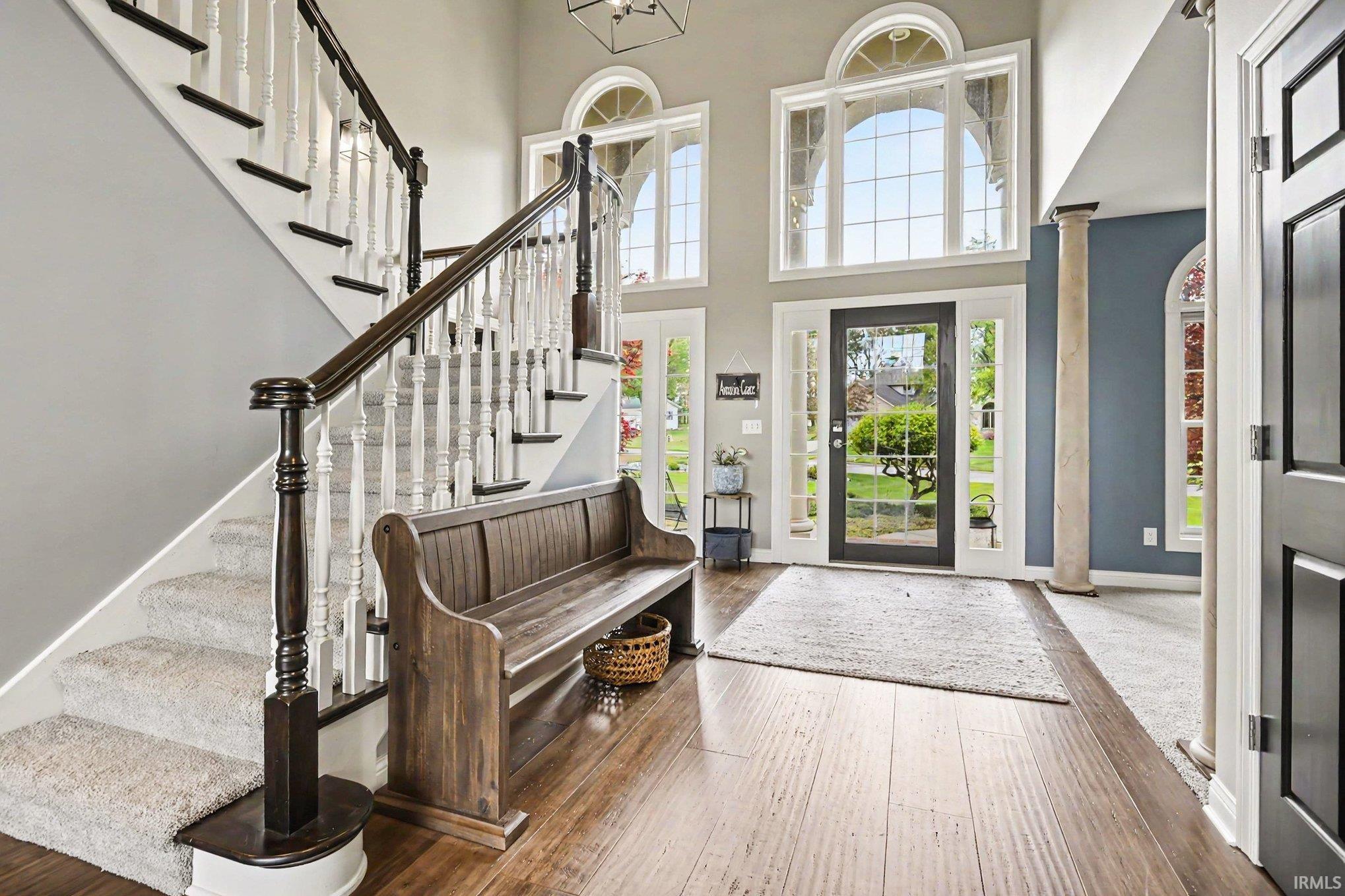 Grand foyer featuring a dual-height ceiling, arched transom windows, wood-finish flooring, a carpeted staircase with white and dark wood banisters, and a decorative column