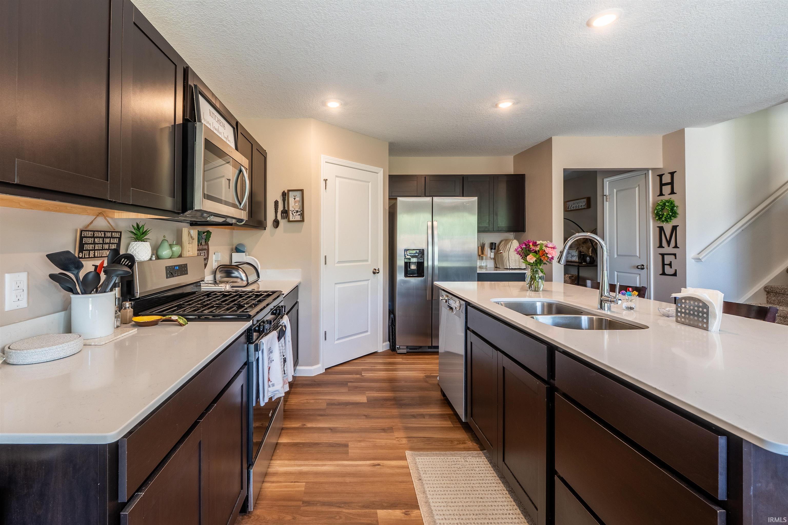 Modern kitchen featuring dark wood-finish cabinetry, light-toned countertops, stainless steel appliances, a center island with a built-in sink, and wood-finish flooring