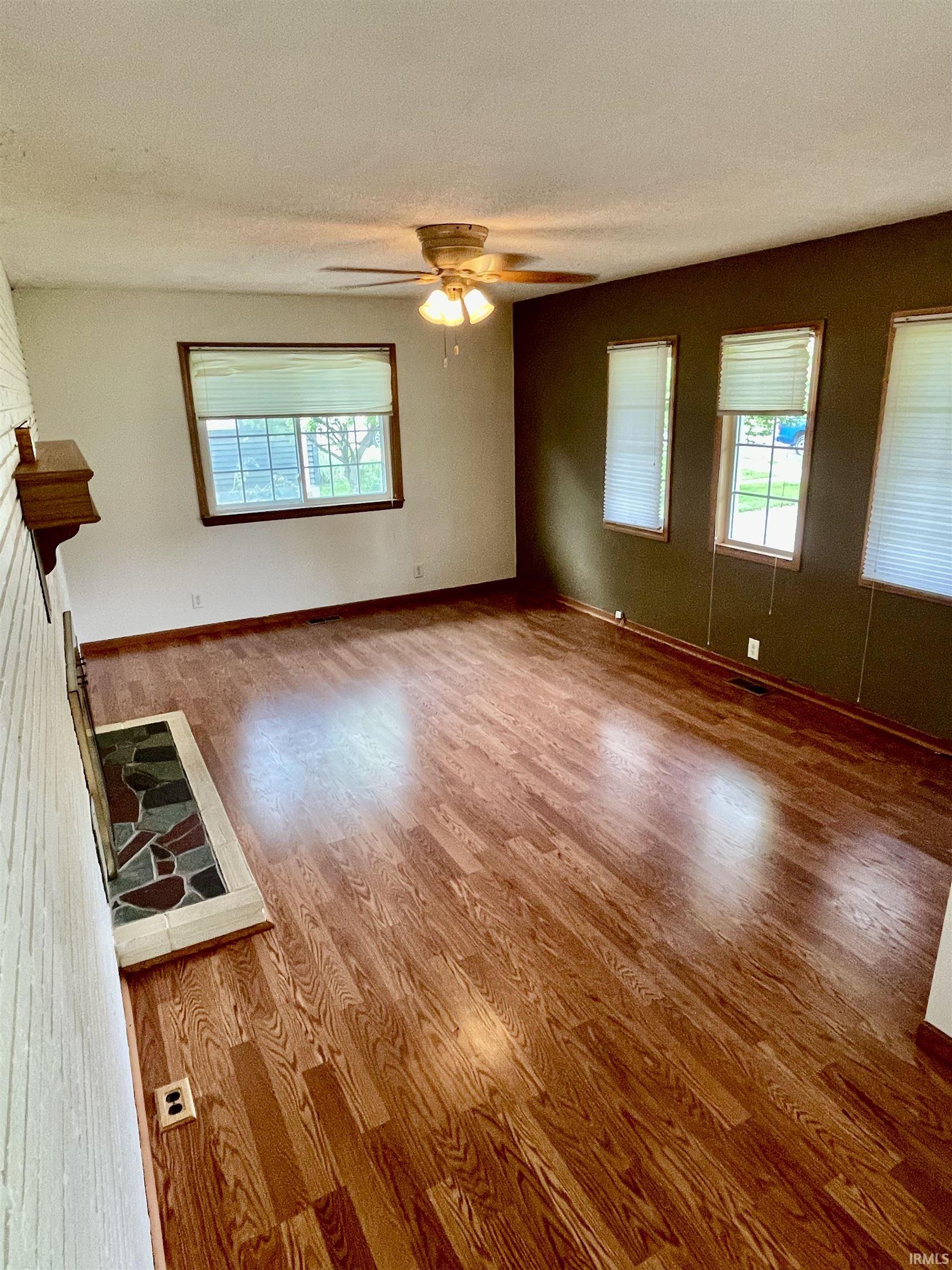 Spacious living area featuring rich wood-finish flooring, a white brick fireplace with a mosaic tile hearth, and multiple windows with blinds