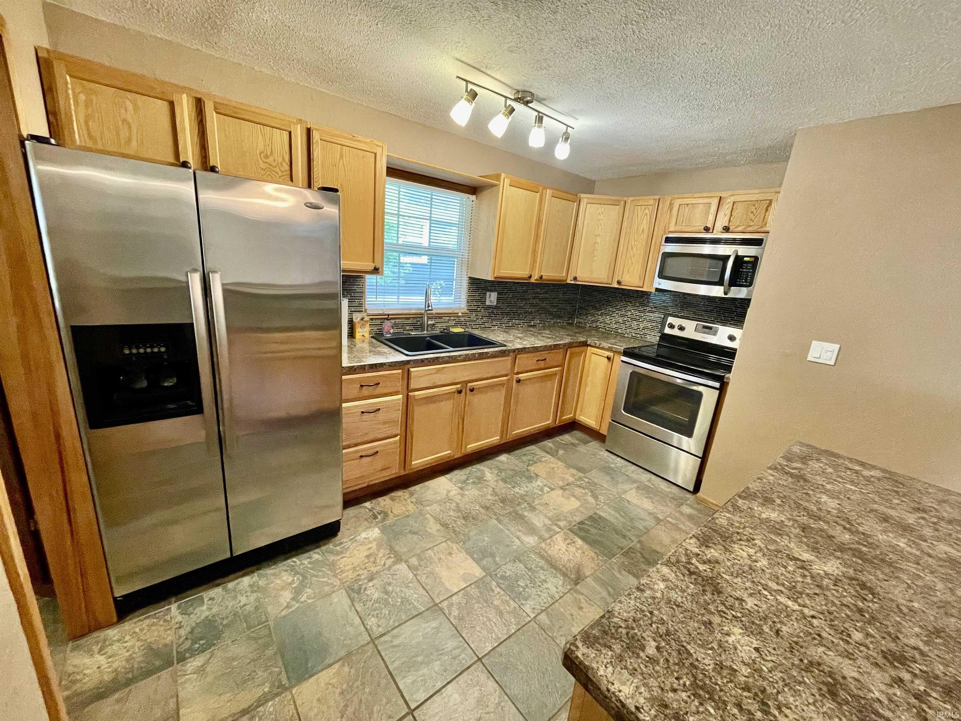 Kitchen featuring wood cabinetry, stainless steel appliances, dark stone-look countertops, and varied-tone square tile flooring