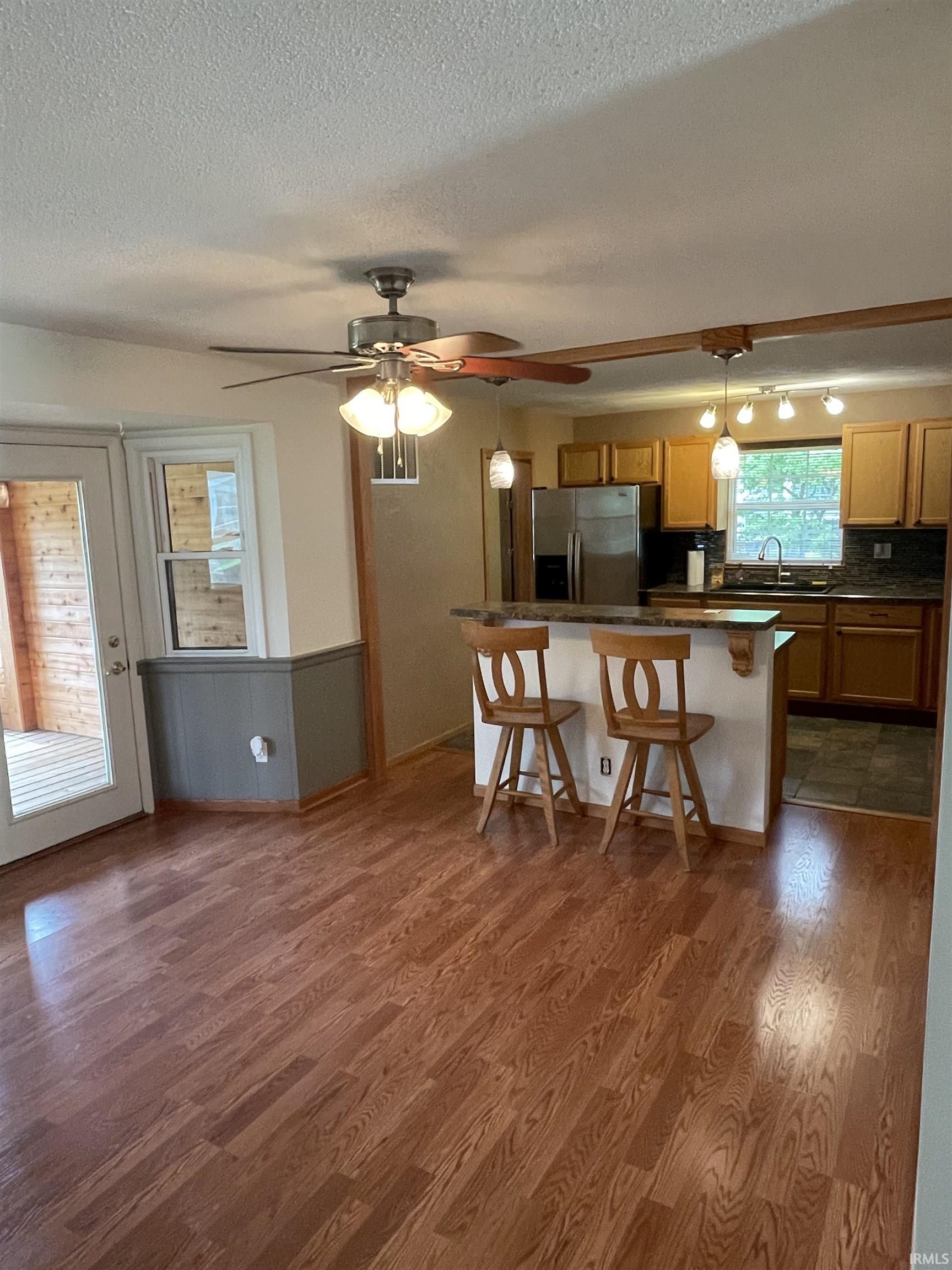 Open-concept kitchen and living area featuring wood-finish flooring, a ceiling fan with integrated lighting, a breakfast bar, stainless steel appliances, and a glass-paneled door