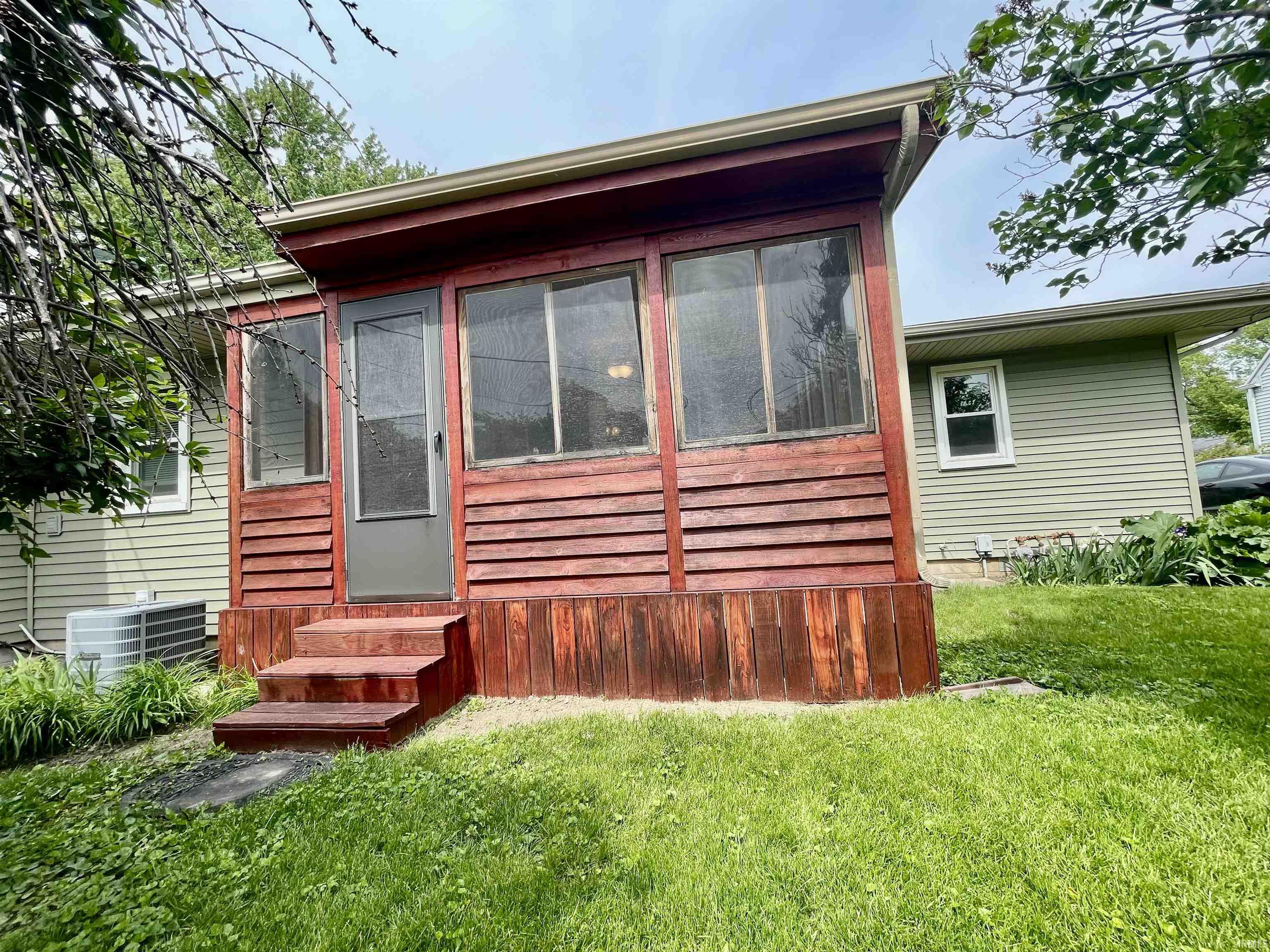 Enclosed porch with wood paneling and multiple windows, featuring a screen door and three steps leading to a grass area