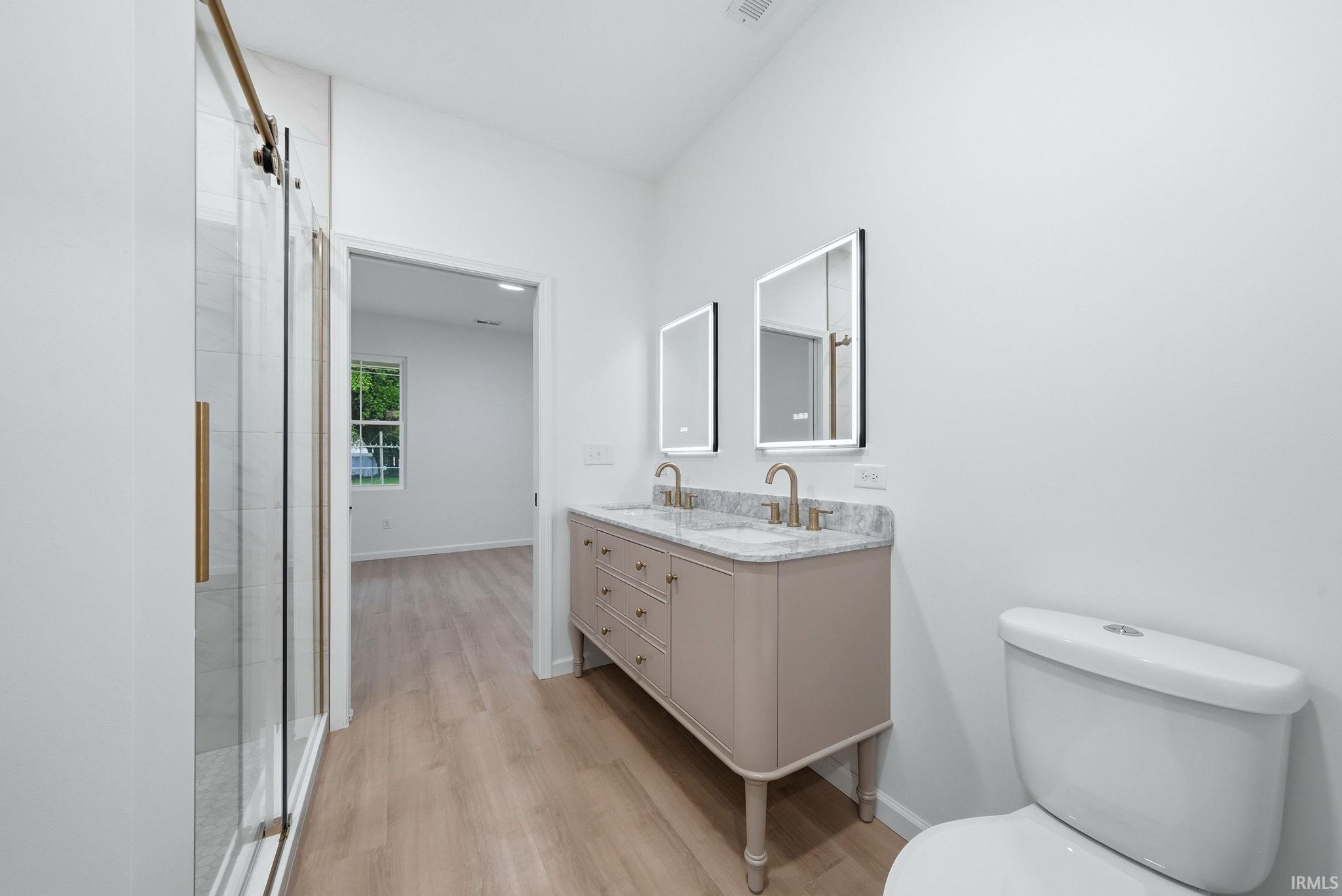 Bright bathroom featuring a dual vanity with a stone countertop, individual illuminated mirrors, and a glass-enclosed shower with a sliding door