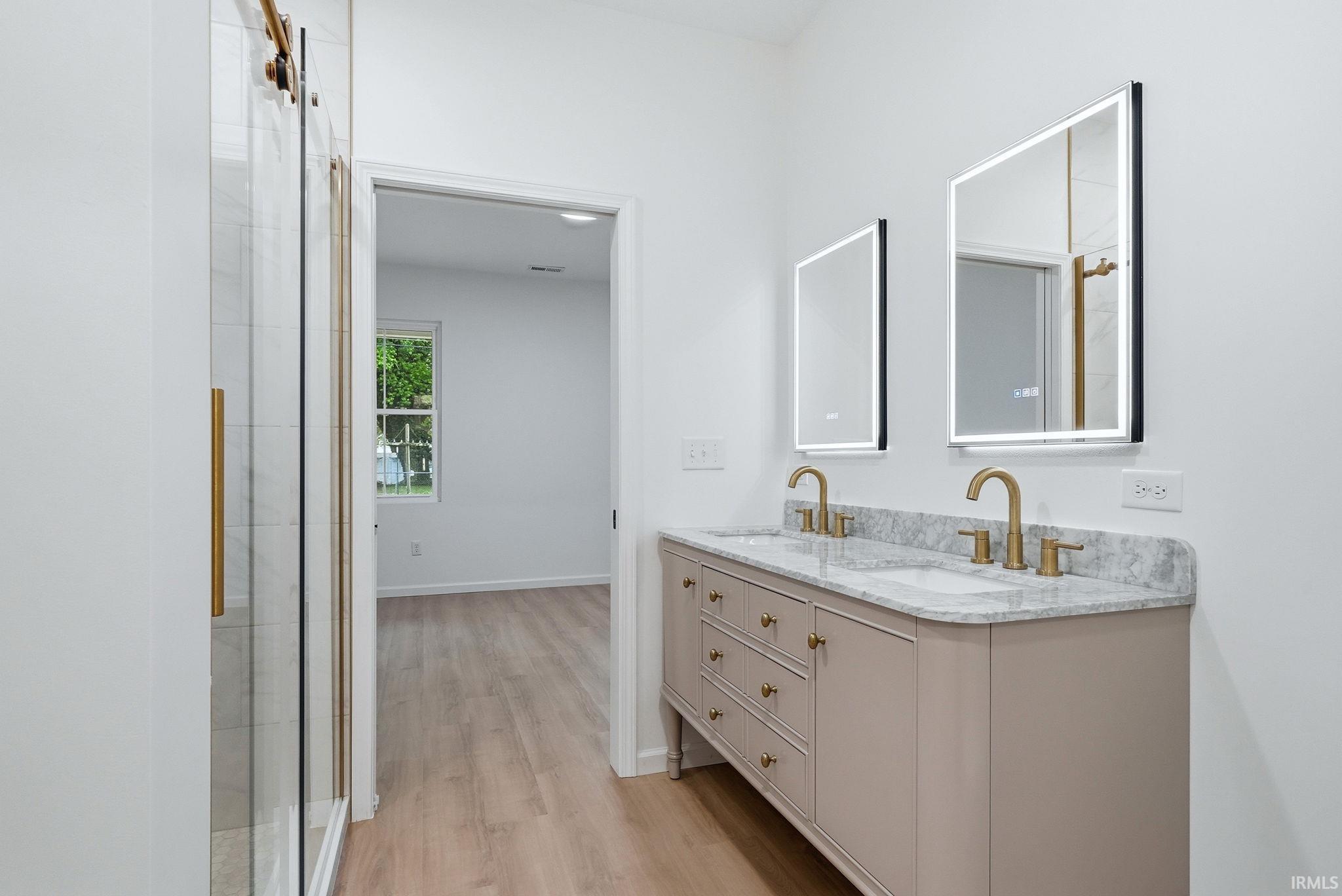 Dual vanity with brushed gold hardware, two illuminated mirrors, and a glass-enclosed shower with gold accents