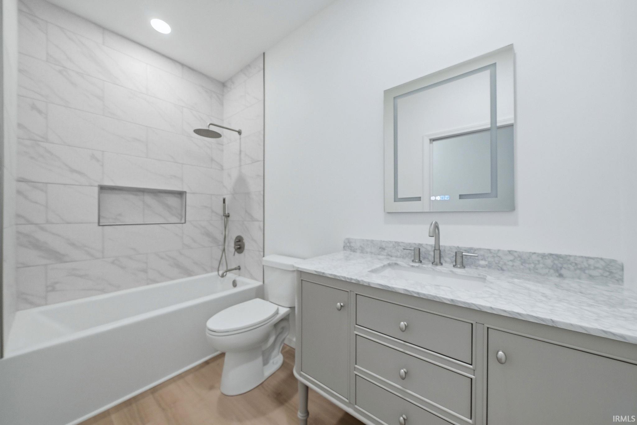 Contemporary bathroom featuring a light gray vanity with a stone countertop, an illuminated mirror, a bathtub with a tiled surround, a recessed shower niche, and wood-finish flooring
