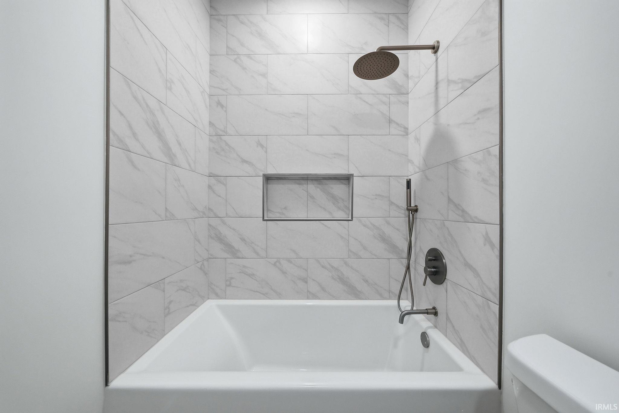 Bathtub and shower combination featuring white marble-look subway tile, a recessed shower niche, and matte bronze fixtures including a rainfall showerhead and handheld sprayer