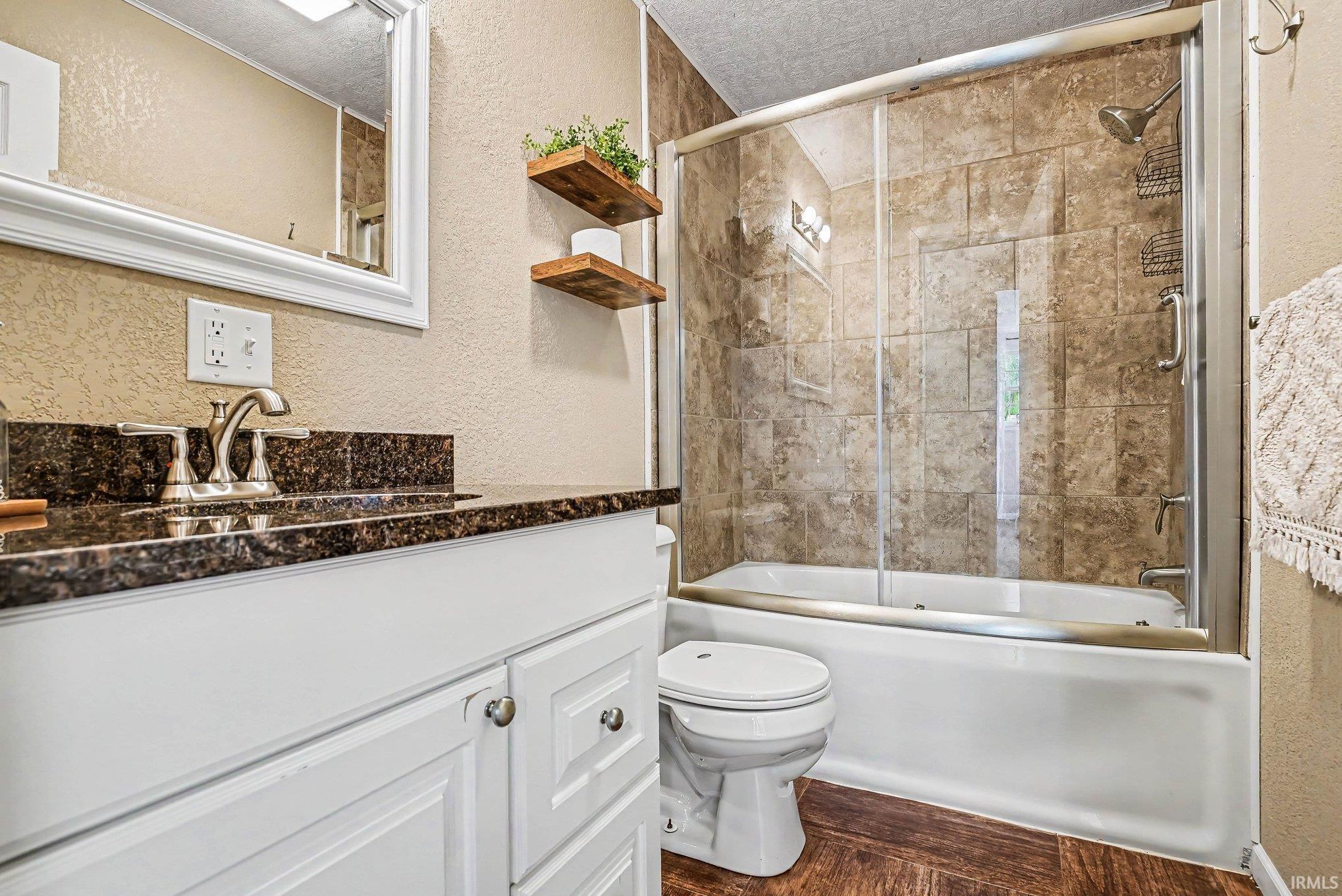 Bathroom featuring a white vanity with a dark countertop and brushed nickel fixtures, a tub and shower combination with glass enclosure and tiled surround, and wood-finish flooring