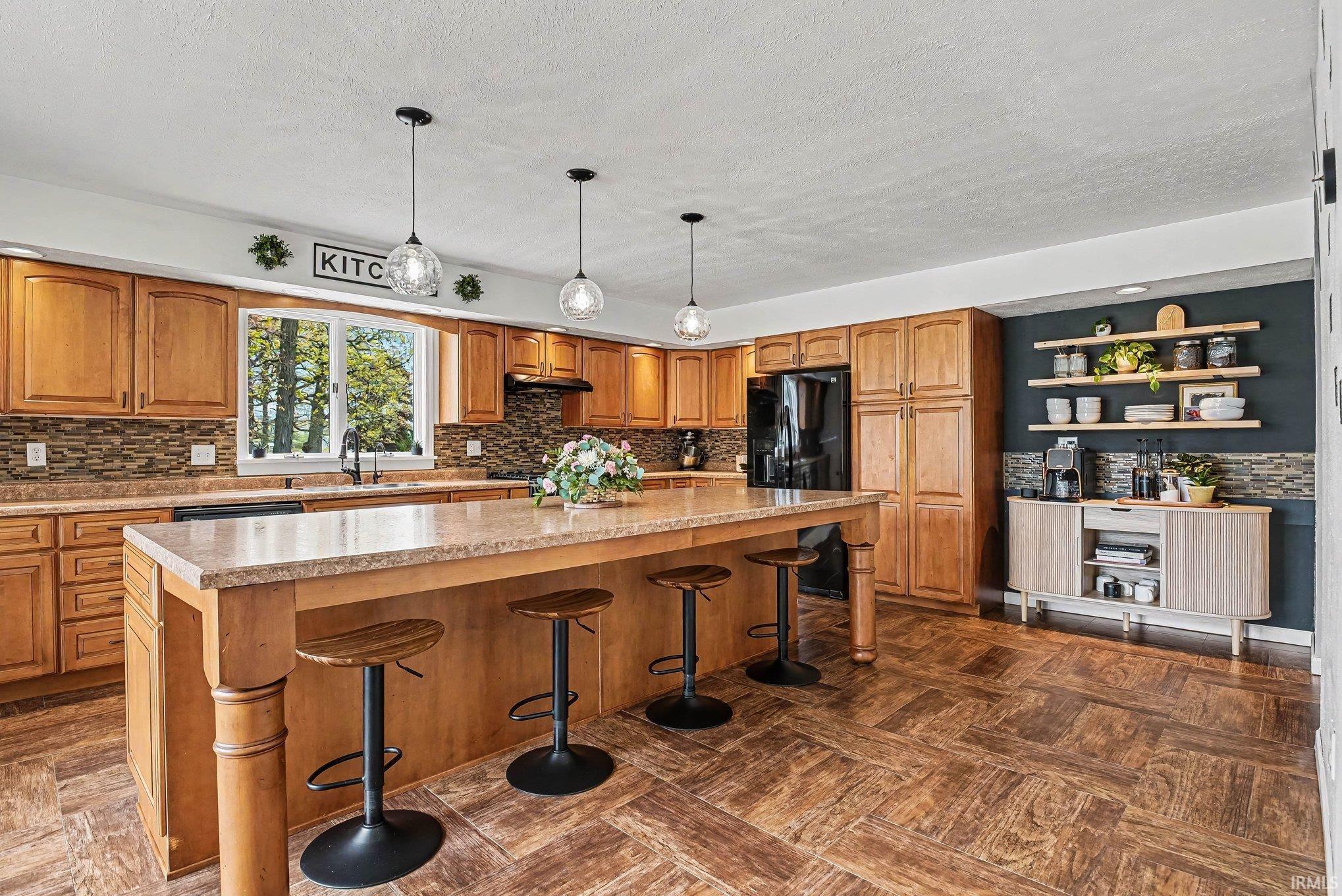 Spacious kitchen featuring a large central island with seating, extensive wood cabinetry, coordinating tile backsplash, dark appliances, and patterned flooring