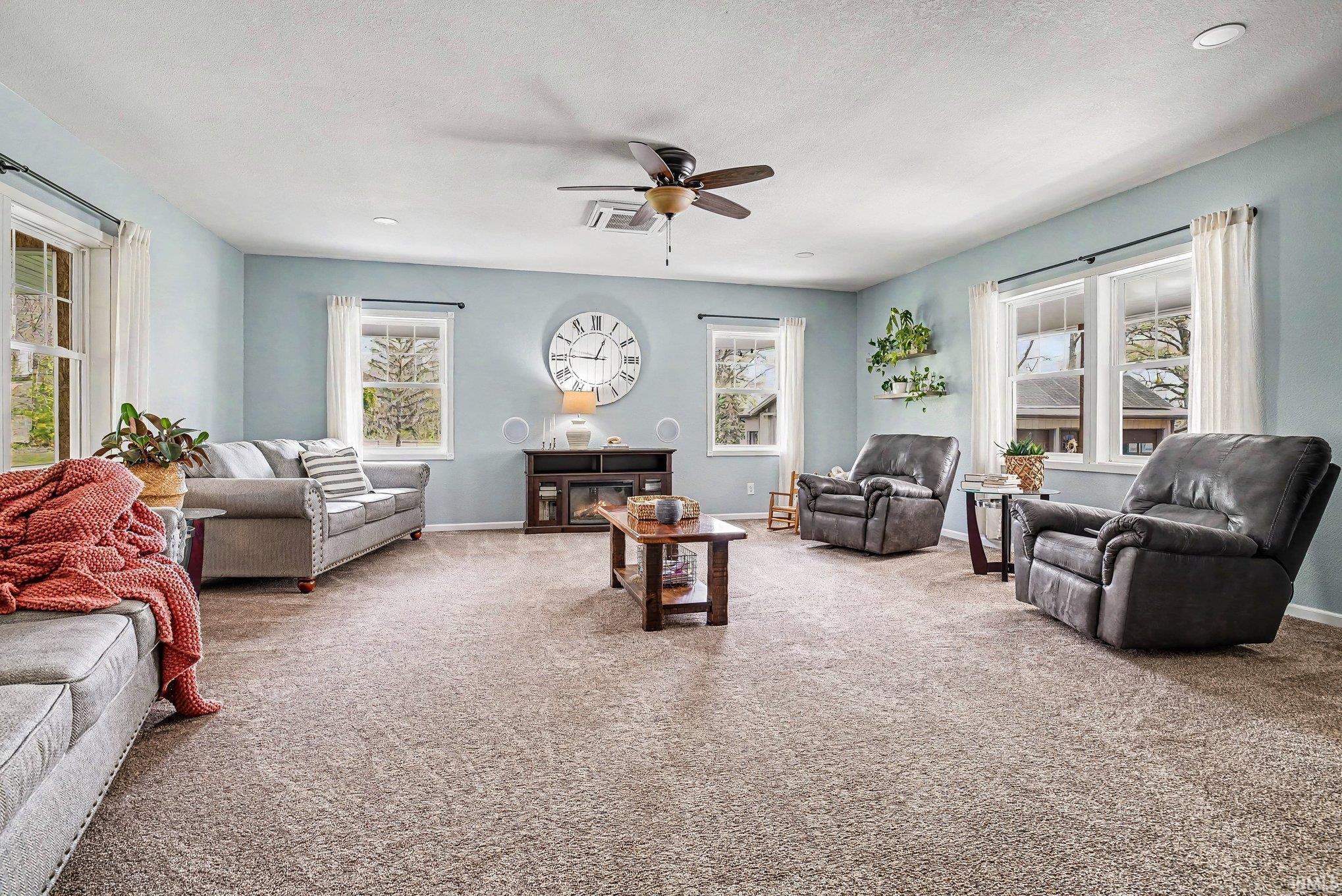 Spacious living area featuring light-colored carpet flooring, multiple windows providing natural illumination, and a ceiling fan with integrated lighting