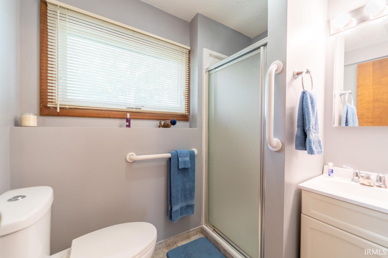 Walk-in shower with frosted glass enclosure, white vanity with integrated sink, and a window featuring wood trim