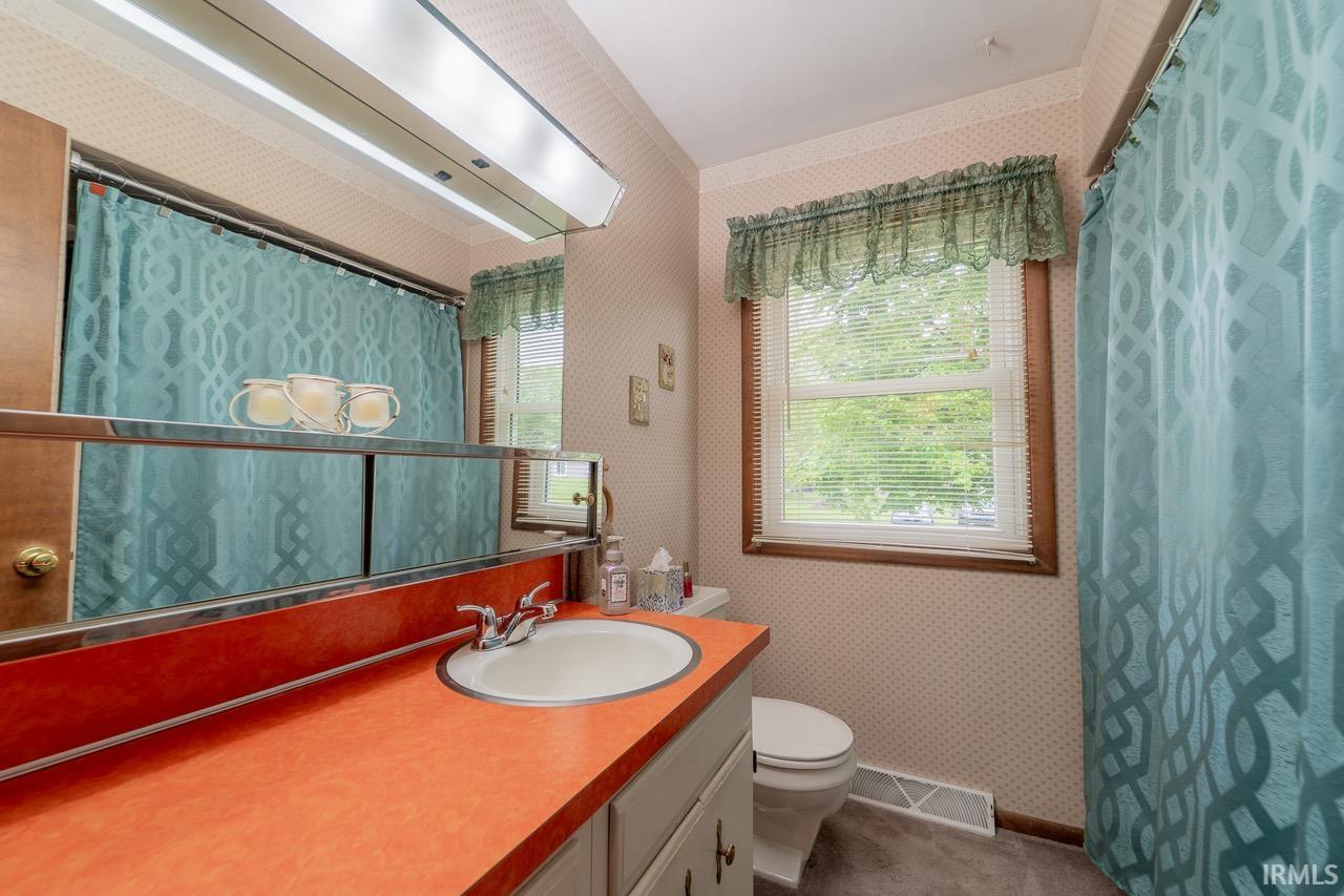 Bathroom featuring a vanity with an integrated sink and orange countertop, a toilet, a window with blinds, and a shower area