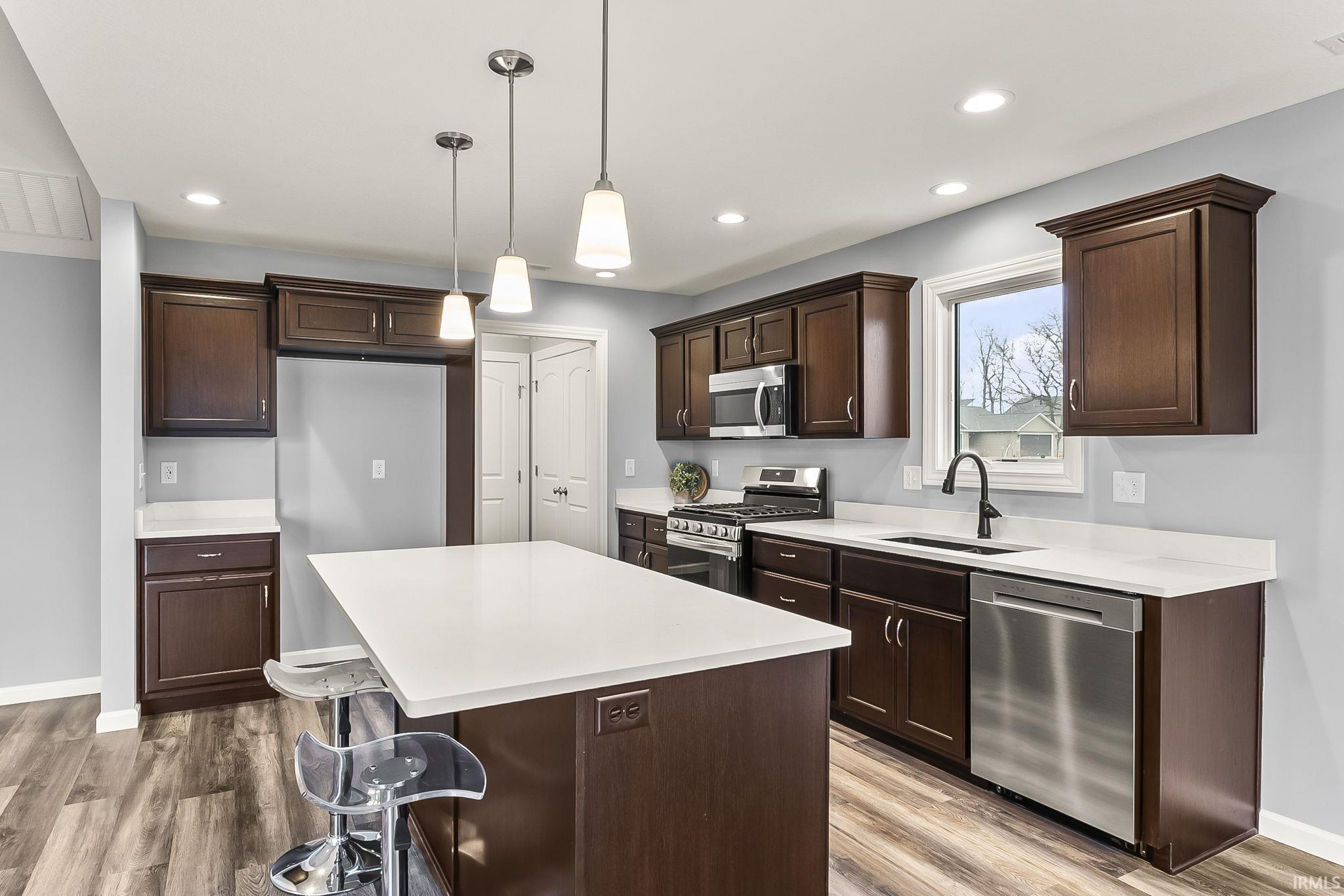 Kitchen featuring wood-finish flooring, dark wood cabinetry, white countertops, stainless steel appliances, and an island with pendant lighting