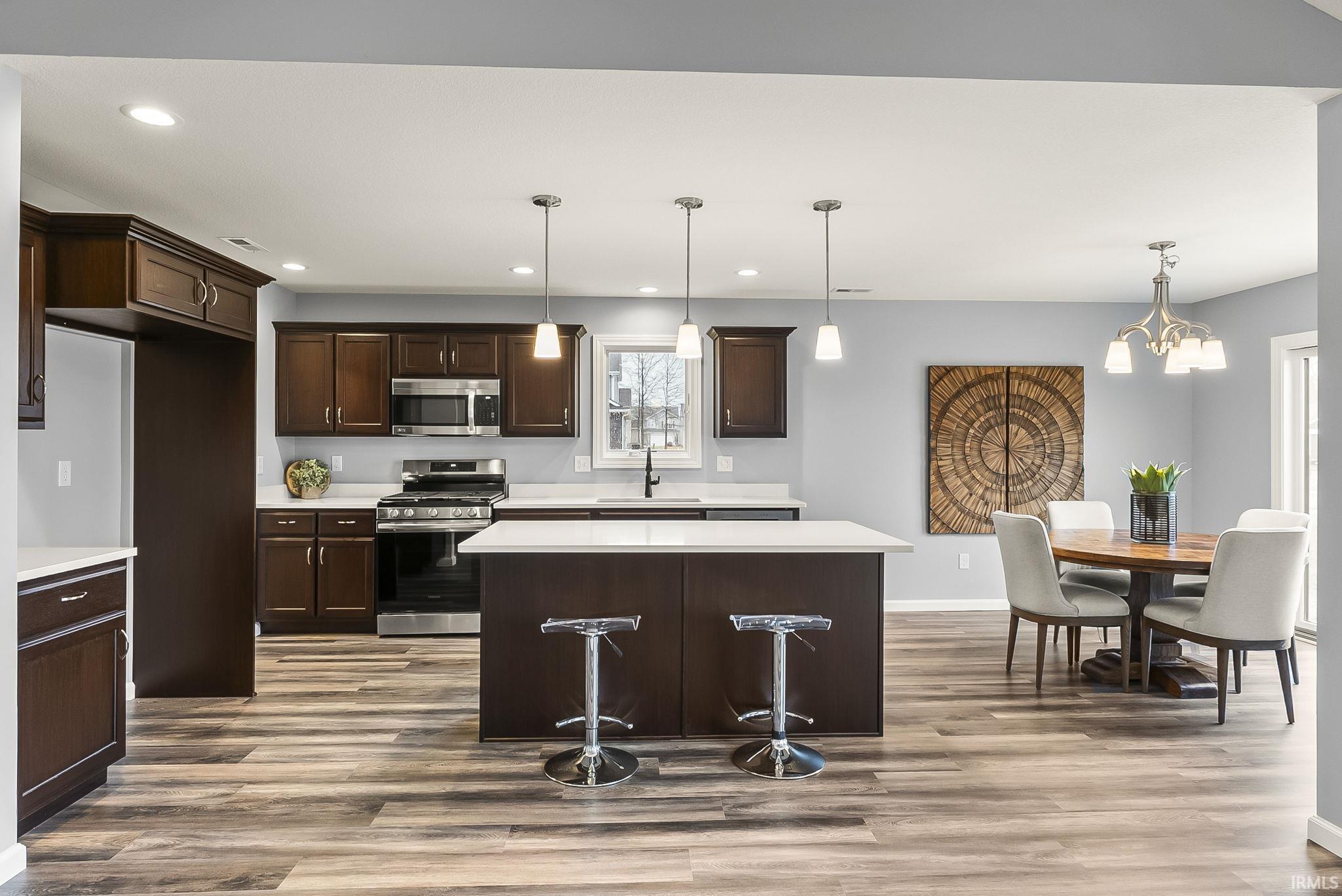 Open-concept kitchen featuring wood-finish flooring, dark wood cabinetry, white countertops, stainless steel appliances, and an island with pendant lighting