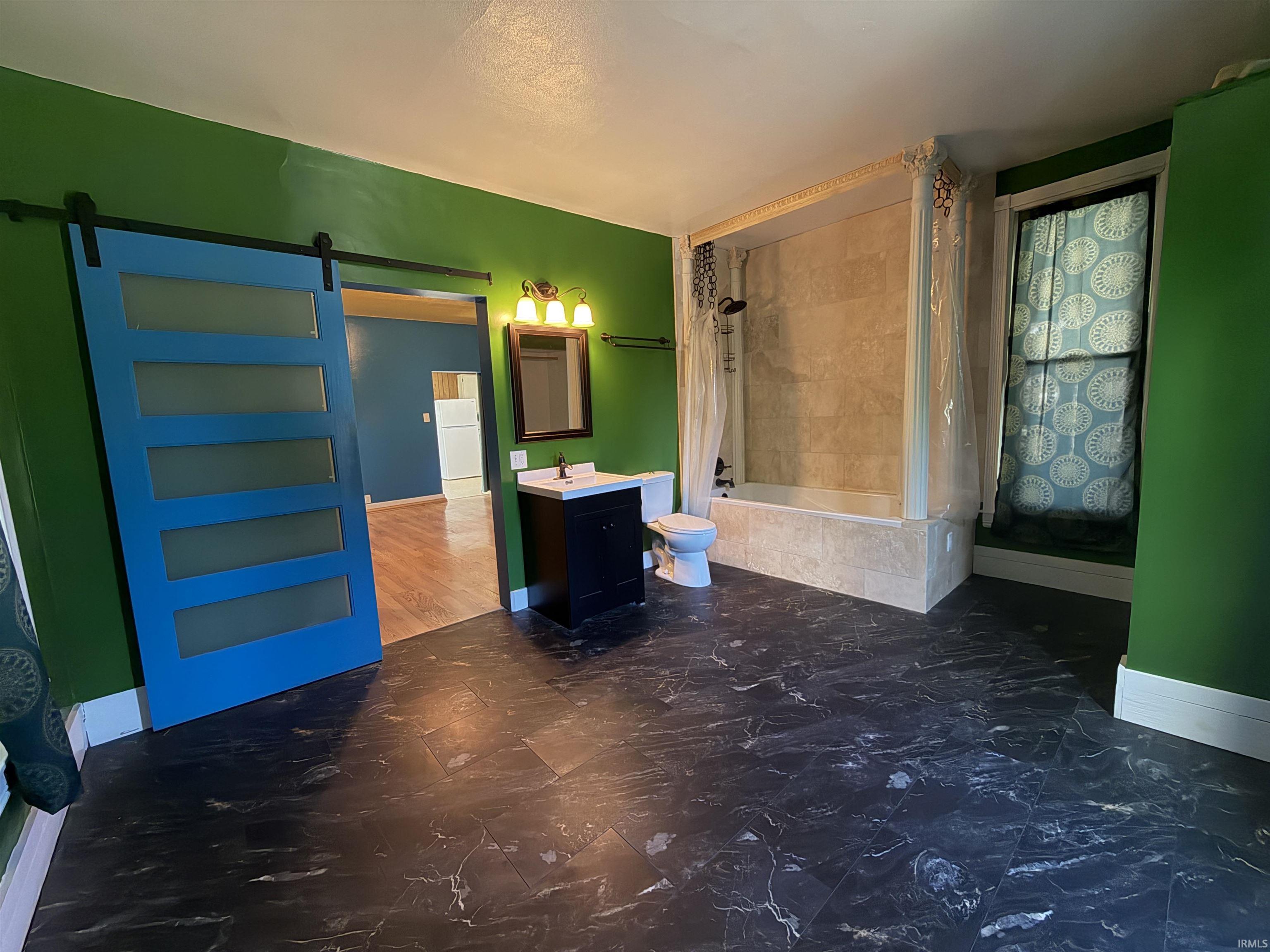 Bathroom featuring black marble-look flooring, a blue sliding barn door with frosted panels, a tub with a tiled surround, a vanity with a white countertop and dark cabinetry, and a mirrored medicine cabinet