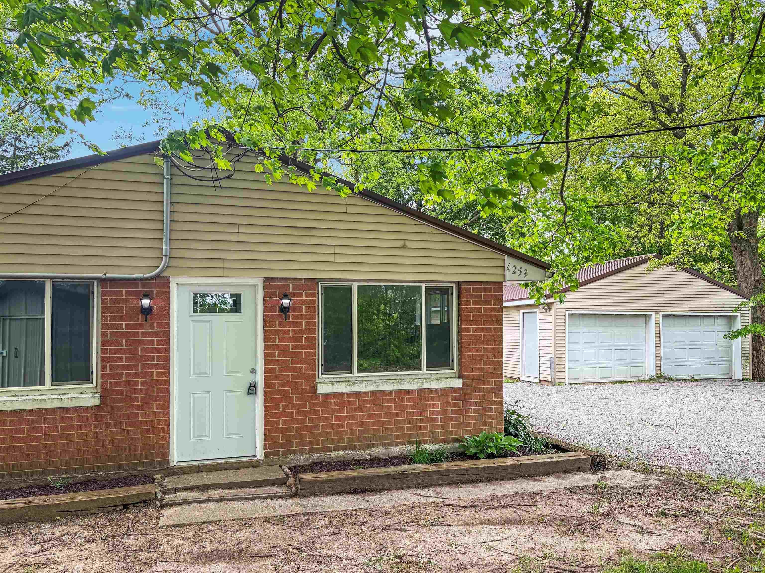 Brick and siding exterior featuring a white entry door with frosted glass insert, two exterior wall sconces, and a gravel driveway leading to a detached two-car garage with two white roll-up doors